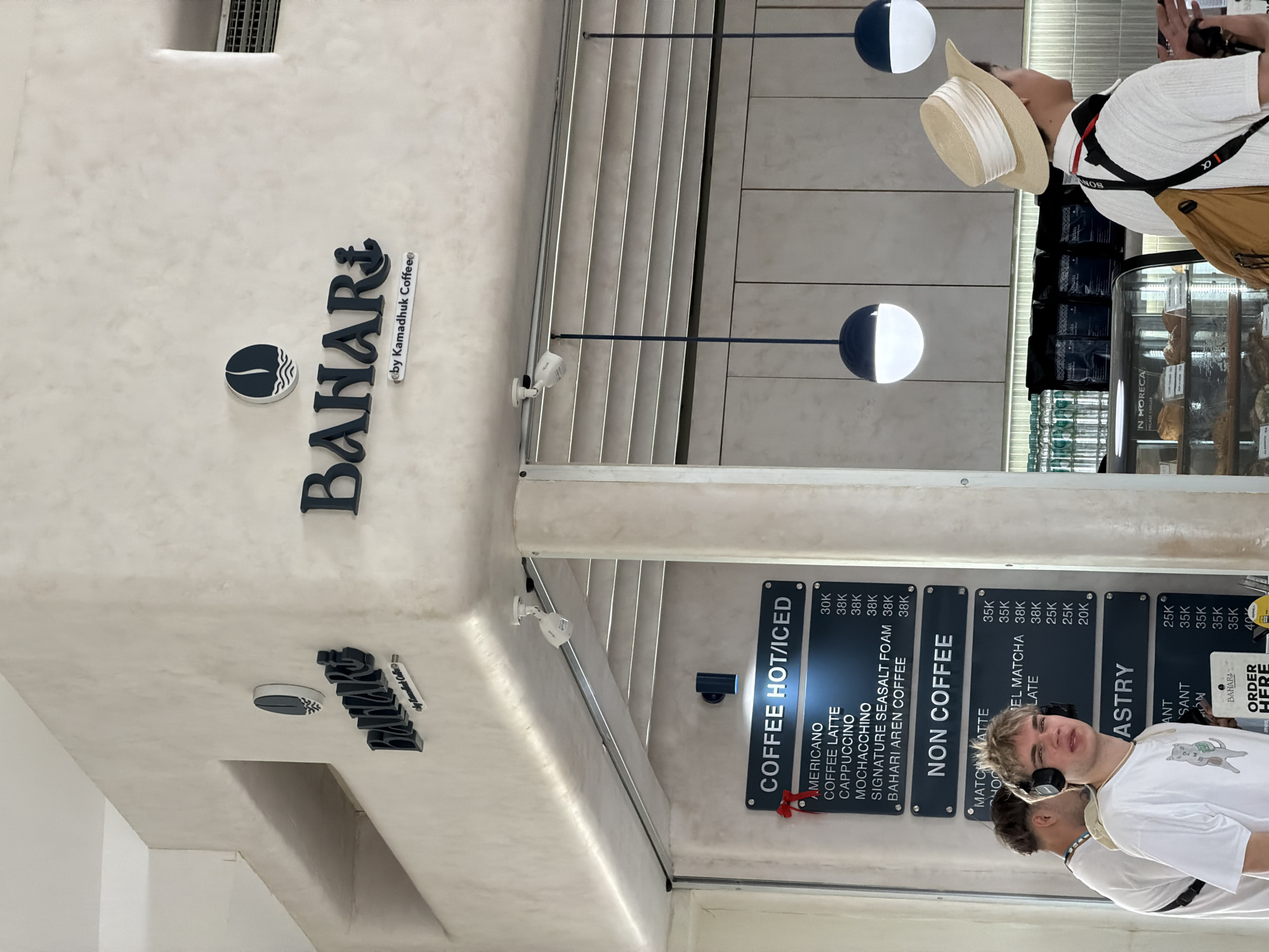 The image shows the entrance of "Bahari by Kamadhuk Coffee" in Denpasar, Indonesia, with the menu displayed on a wall-mounted board. The menu includes "Coffee Hot/Iced" options such as Americano, Coffee Latte, Cappuccino, and signature drinks, as well as "Non Coffee" options and "Pastry". A sign points to "Order Here". Two people are visible in the frame. One person wears a sun hat and is taking a photo, and another person, wearing headphones, is standing near the menu. The architectural design includes a light-colored ceiling and walls with modern lighting fixtures.