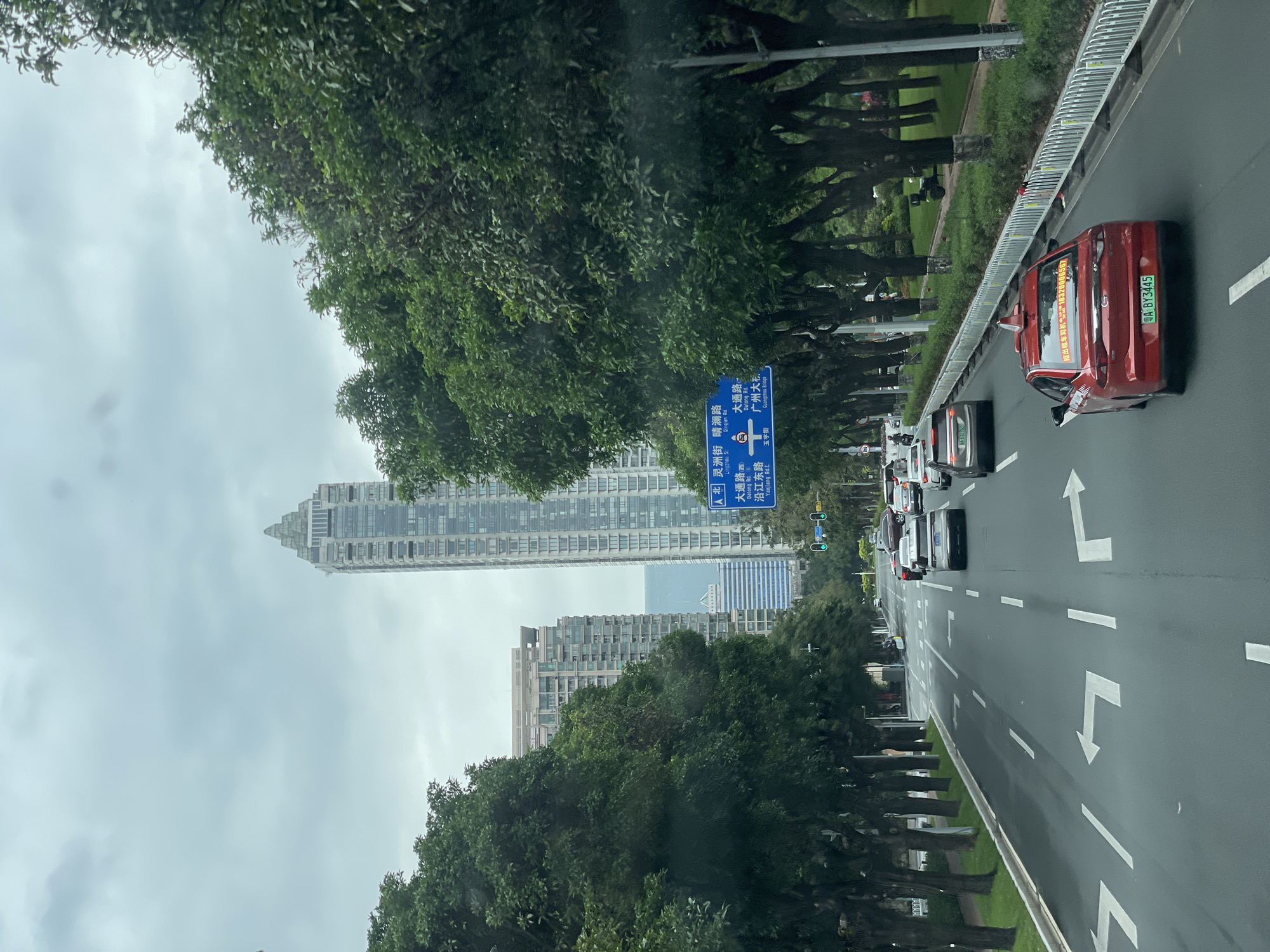 The image shows a street scene in Yuexiu District, Guangzhou, China. Cars are driving on a multi-lane road marked with white arrows indicating direction. Trees line the side of the road, separating the road from the sidewalk. A blue street sign indicates "Lingzhou Street," "Datong Road," and "Yanjiang East Road." In the background, tall skyscrapers are visible through the trees. A red taxi with license plate "A BY3445" is in the right lane. The sky is overcast.