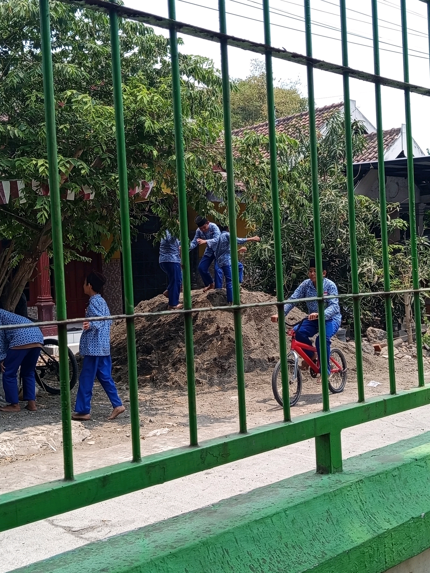 In Randubango, Indonesia, several boys in blue patterned uniforms and dark caps are seen outdoors, some interacting with a dirt mound, while one rides a red bicycle. The scene is partially obstructed by green metal bars. The environment includes trees with green foliage, a building with a tiled roof, and a dirt pathway. The overall impression is a candid snapshot of children engaged in playful activities in a semi-urban or rural setting.