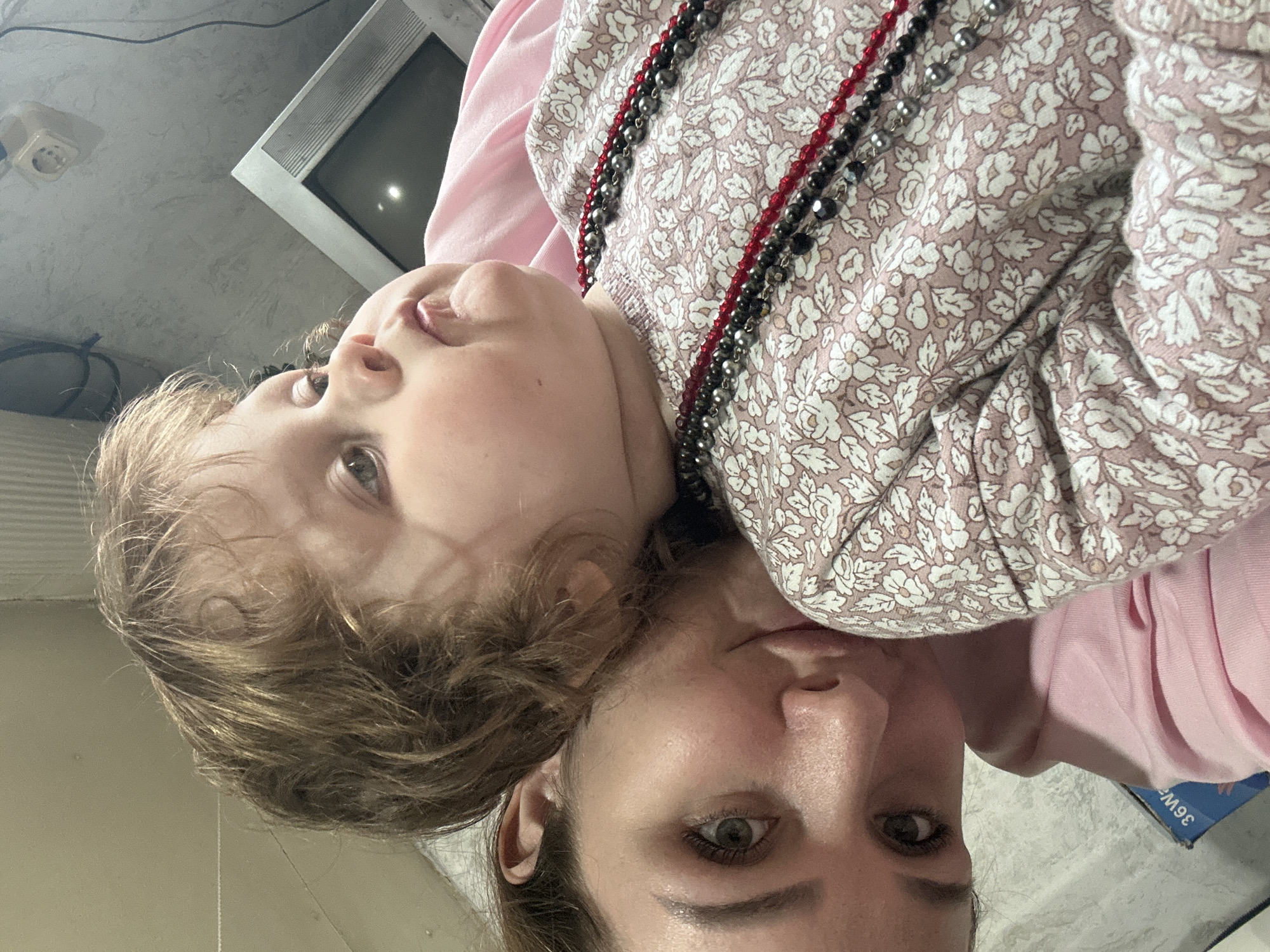 A close-up, face-on shot features a woman and a young child together. The child, wearing a floral patterned top and layered necklaces of red and black beads, rests against the woman, who appears to be holding the camera. The woman has fair skin and is looking into the camera. An older television set is visible in the background, along with electrical outlets on the wall. The overall setting appears to be indoors. The city of Chaikovsky, Russia, is not discernible from the image.