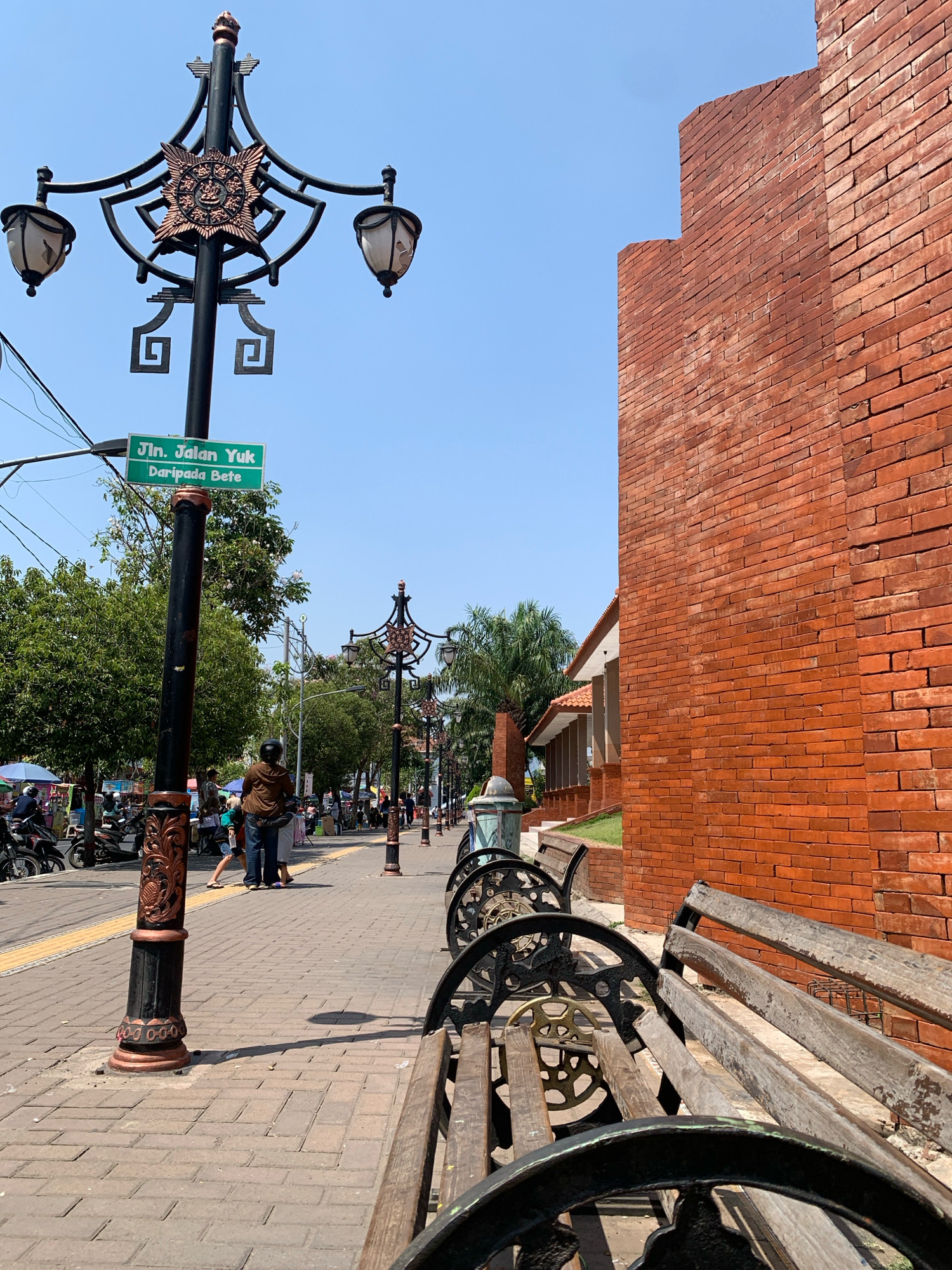 The image depicts a street scene in Mojokerto, Indonesia, characterized by a clear blue sky and bright sunlight. In the foreground, several empty wooden benches with ornate metal frames are visible, positioned on a paved pedestrian walkway. To the left, a lamppost with intricate decorative elements and two lamps stands prominently, bearing a green street sign that reads "Jln. Jalan Yuk Daripada Bete." Beyond the lamppost, a busy street is visible with motorbikes, pedestrians, and market stalls. A tall brick wall, characteristic of Indonesian architecture, forms a backdrop on the right side of the image, its warm red tones contrasting with the blue sky. The overall atmosphere suggests a vibrant and bustling urban environment on a sunny day.