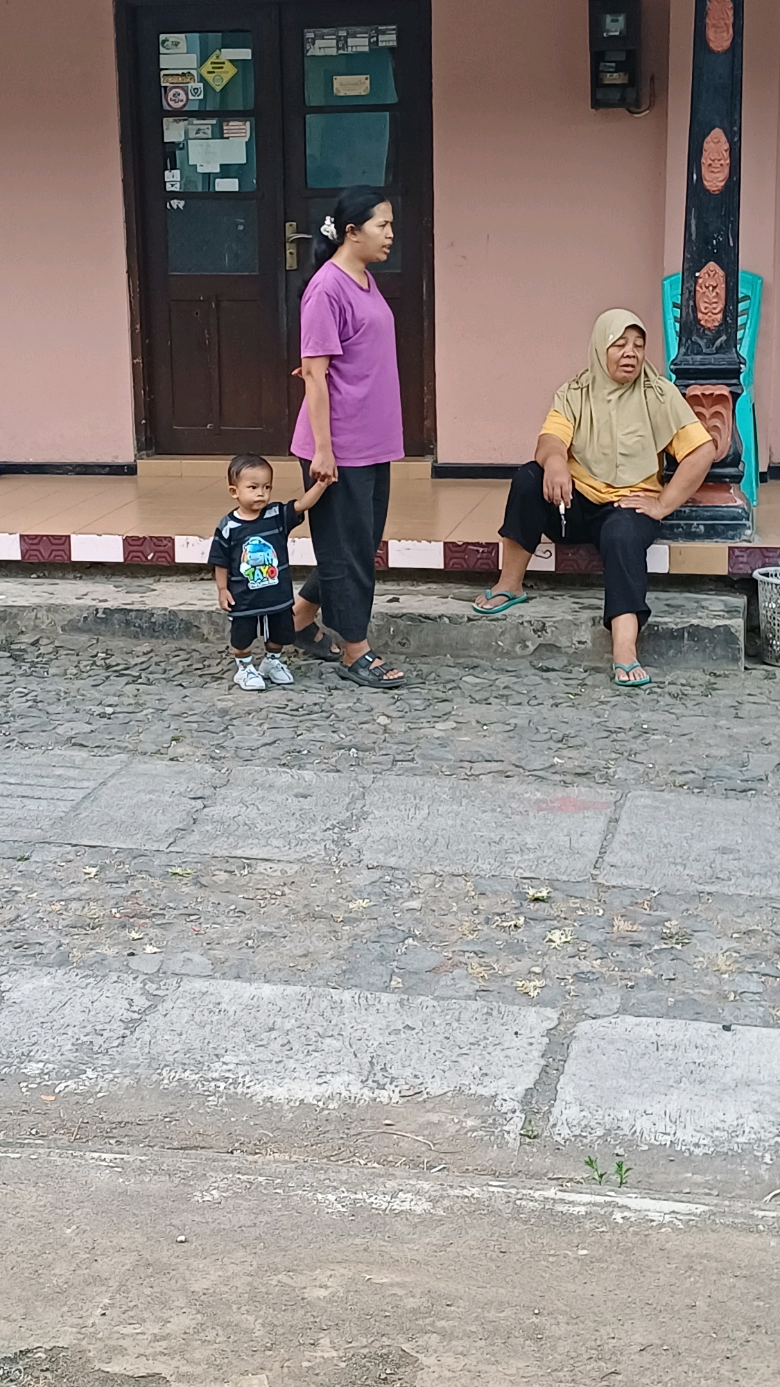 Three individuals are present outside a building. On the left, a young child, wearing a black t-shirt with a graphic print and shorts, stands holding the hand of an adult woman dressed in a purple t-shirt and dark pants. To the right, an adult woman wearing a light brown headscarf, a yellow t-shirt, and dark pants is seated on a step. The ground in front of the building is paved with stones and concrete. The building facade is light pink with a dark brown door. A black pillar with red ornamentation is visible on the right side of the image. The scene appears to be in Wonokerso, Indonesia.