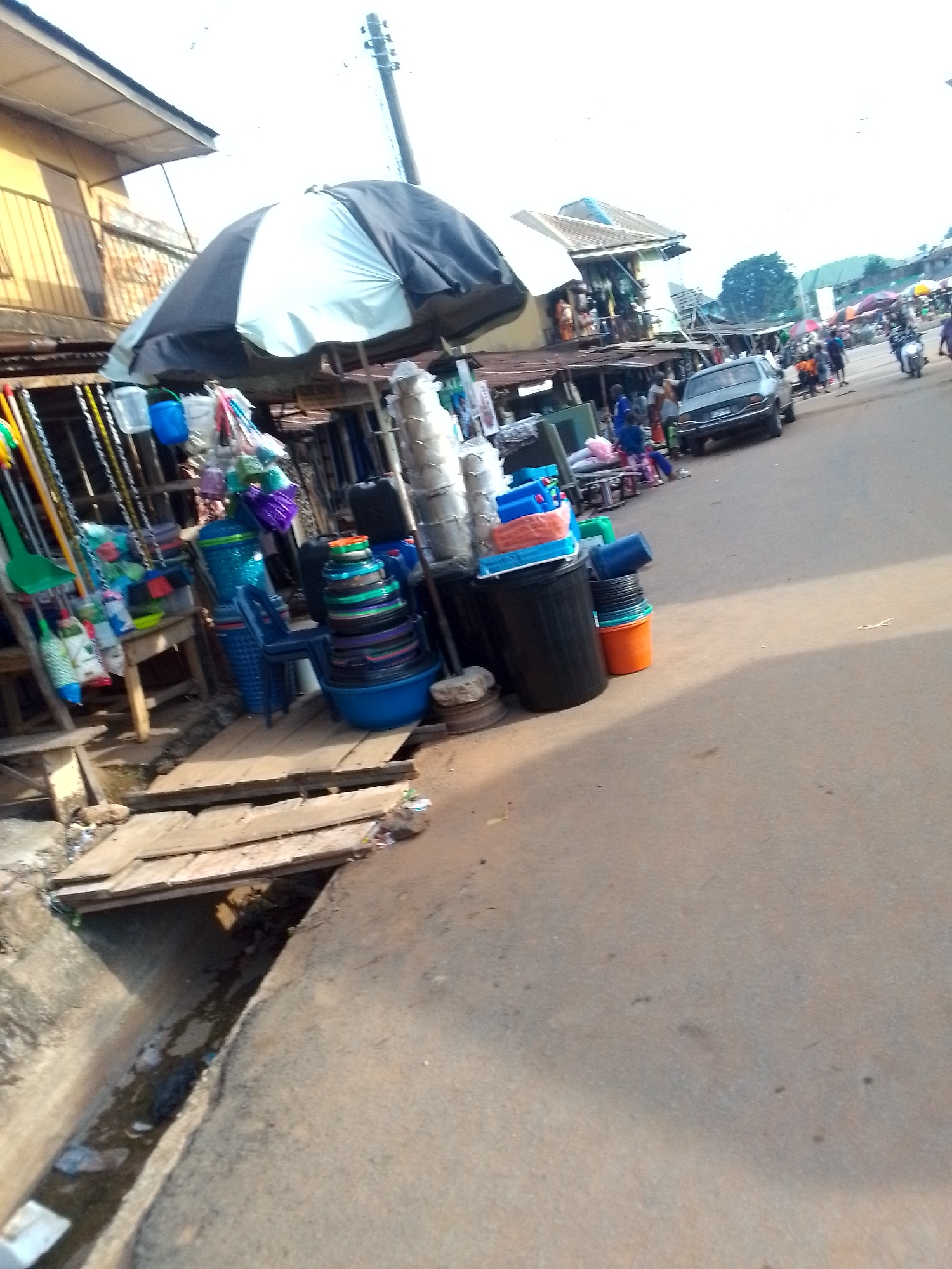 The image depicts a street scene in Orlu, Nigeria. A vendor's stall occupies the left side of the frame, displaying various household items, including buckets, basins, brooms, and plastic containers, under a large umbrella. A makeshift wooden bridge covers a drainage ditch in the foreground, partially filled with debris. The street slopes upwards, revealing a car parked along the roadside and other people on the street, including individuals on a motorcycle. Buildings line the street, with visible commercial activity.