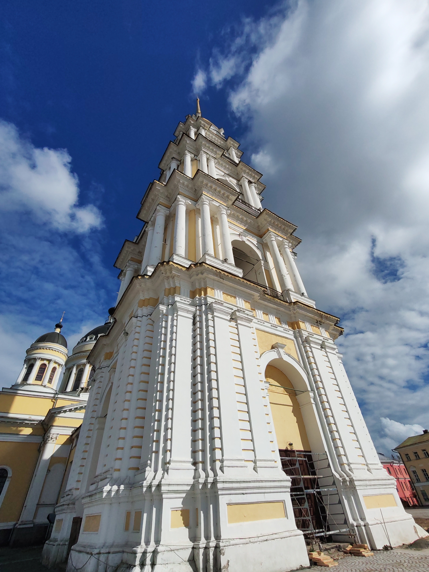 A low-angle, wide-shot photograph captures the striking architecture of a white and yellow Orthodox church bell tower and adjoining domes against a bright blue sky with scattered white clouds. The bell tower is the primary subject, rising majestically with multiple tiers, Corinthian columns, and decorative cornices. Its base features a large arched entryway, partially obscured by scaffolding and construction materials. To the left, two onion domes of the church are visible, topped with crosses. The building's facade is adorned with intricate detailing, including repetitive rectangular indentations along the tower's body and ornate gold trim. The scene is set in Bol'shie Mkhi, Russia. In the background, the corner of a building with a red facade and another with a yellow facade can be seen. The ground in the foreground is paved with cobblestones. No people are present in the image.