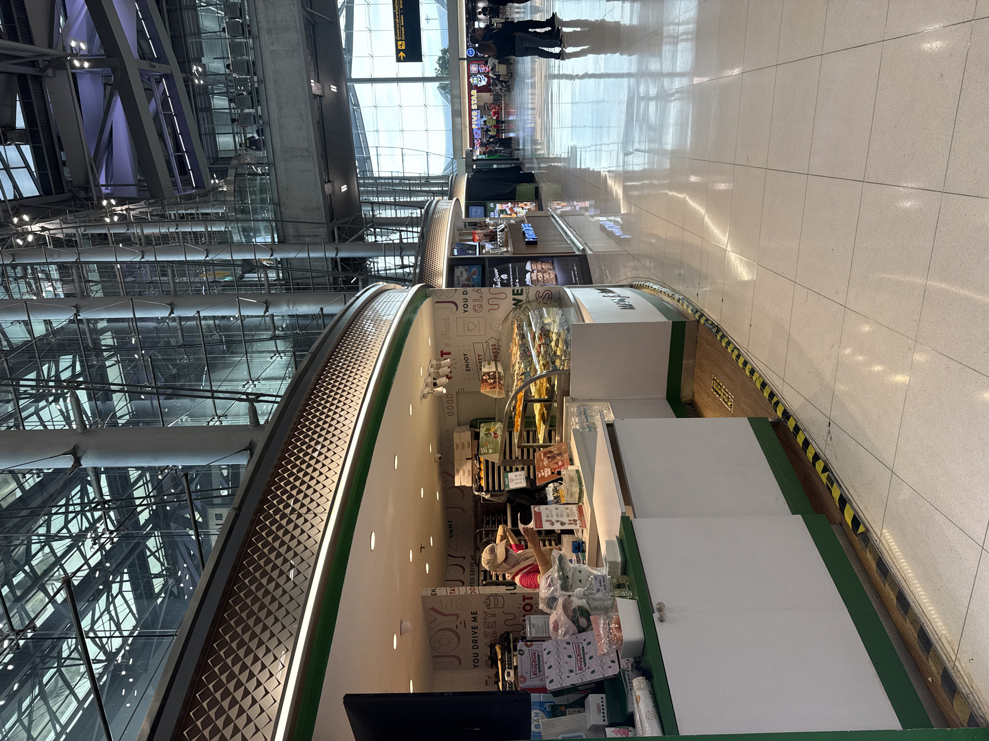 The image depicts an interior view of an airport terminal in Nong Prue Subdistrict, Thailand. A Krispy Kreme donut shop is visible on the left, with a woman wearing a mask and a cap behind the counter, appearing to serve customers or prepare orders. Various donut boxes and displays are present. Adjacent to the Krispy Kreme, a P.Thong Coffee shop is also visible. The terminal's architecture features a modern design with a glass ceiling and large structural pillars. In the background, other shops and travelers are seen, with signage indicating "Domestic Arrivals." The flooring is tiled and reflects the overhead lighting. The scene captures a moment of daily activity within a busy transportation hub.