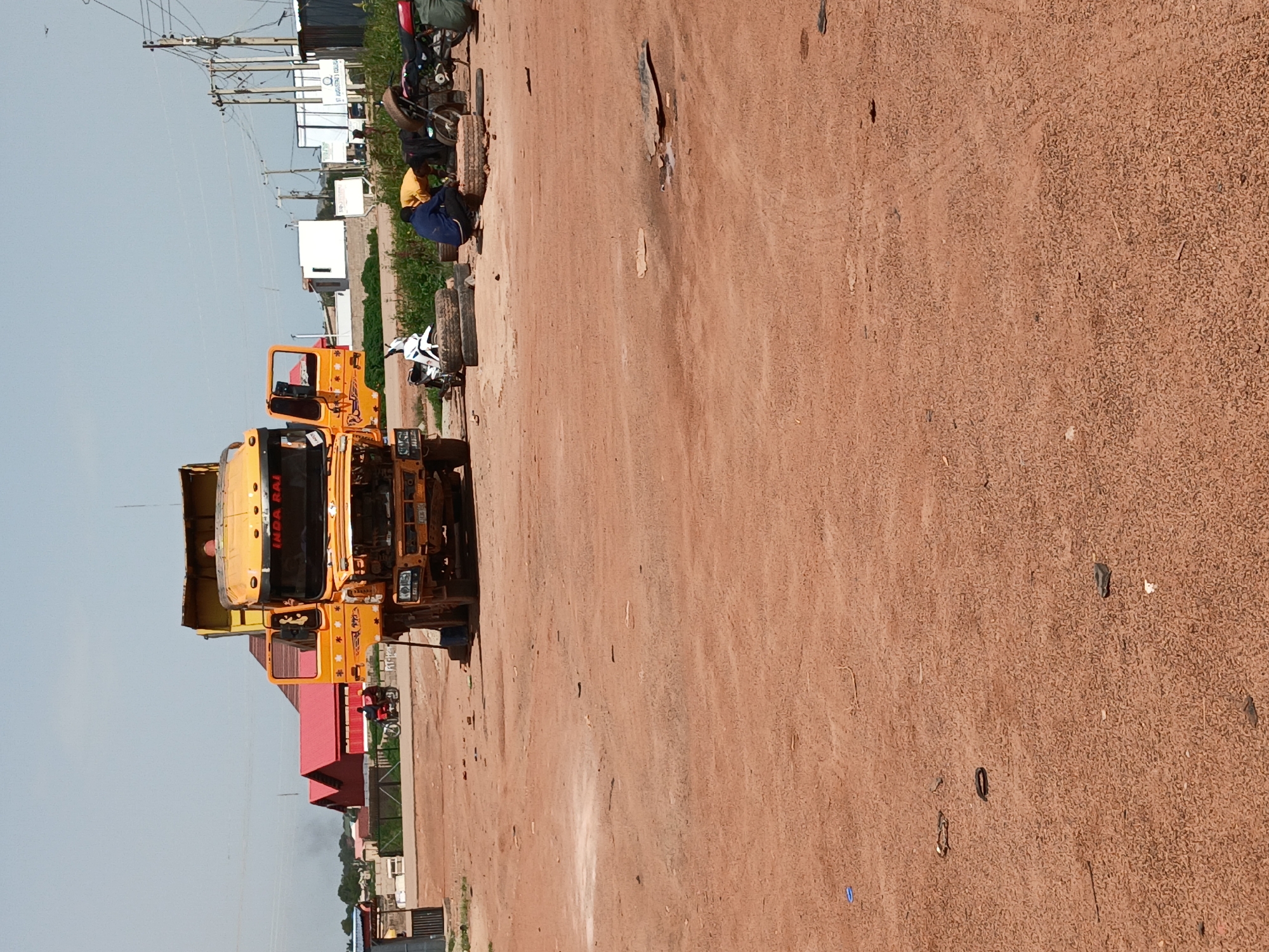 In Gura Top, Nigeria, a yellow dump truck is parked on a dirt lot, its engine compartment open and hood raised. The truck is the central focus, with its cab doors ajar. To the right of the truck, two individuals are engaged in activity. One person, wearing a yellow shirt and blue pants, is kneeling near a stack of tires and appears to be working on a motorcycle. Another person, wearing a blue jacket and dark pants, is also near the tires and motorcycle. A white motorcycle is parked to the left of the truck, and a red motorcycle is visible further to the right. The environment is dusty, with reddish-brown dirt covering the ground. In the background, several buildings with red roofs are visible, along with utility poles and wires, suggesting an urban or semi-urban setting. The sky is clear and bright, indicating daytime.