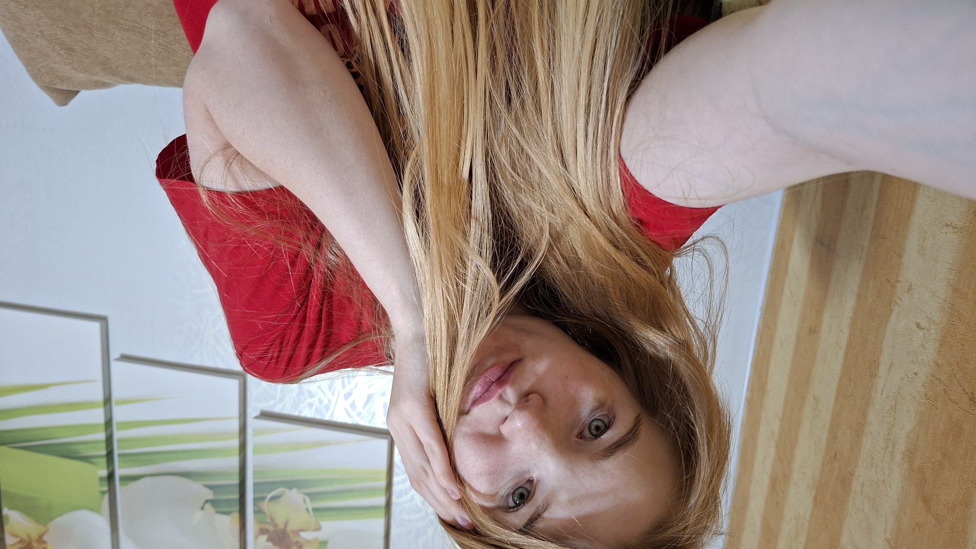 The image shows a close-up of a young woman with long, blonde hair and green eyes. She is wearing a red t-shirt and her left arm is extended towards the camera, as if taking a selfie. Her right hand is touching her face. In the background, there is a wall with a light-colored textured wallpaper and a framed picture with floral elements. To the left of the woman, there is a wall with vertical wooden paneling. The overall impression is of a casual, indoor setting, possibly a bedroom or living room. There is no identifiable information about the location being in Большой Букор, Russia.