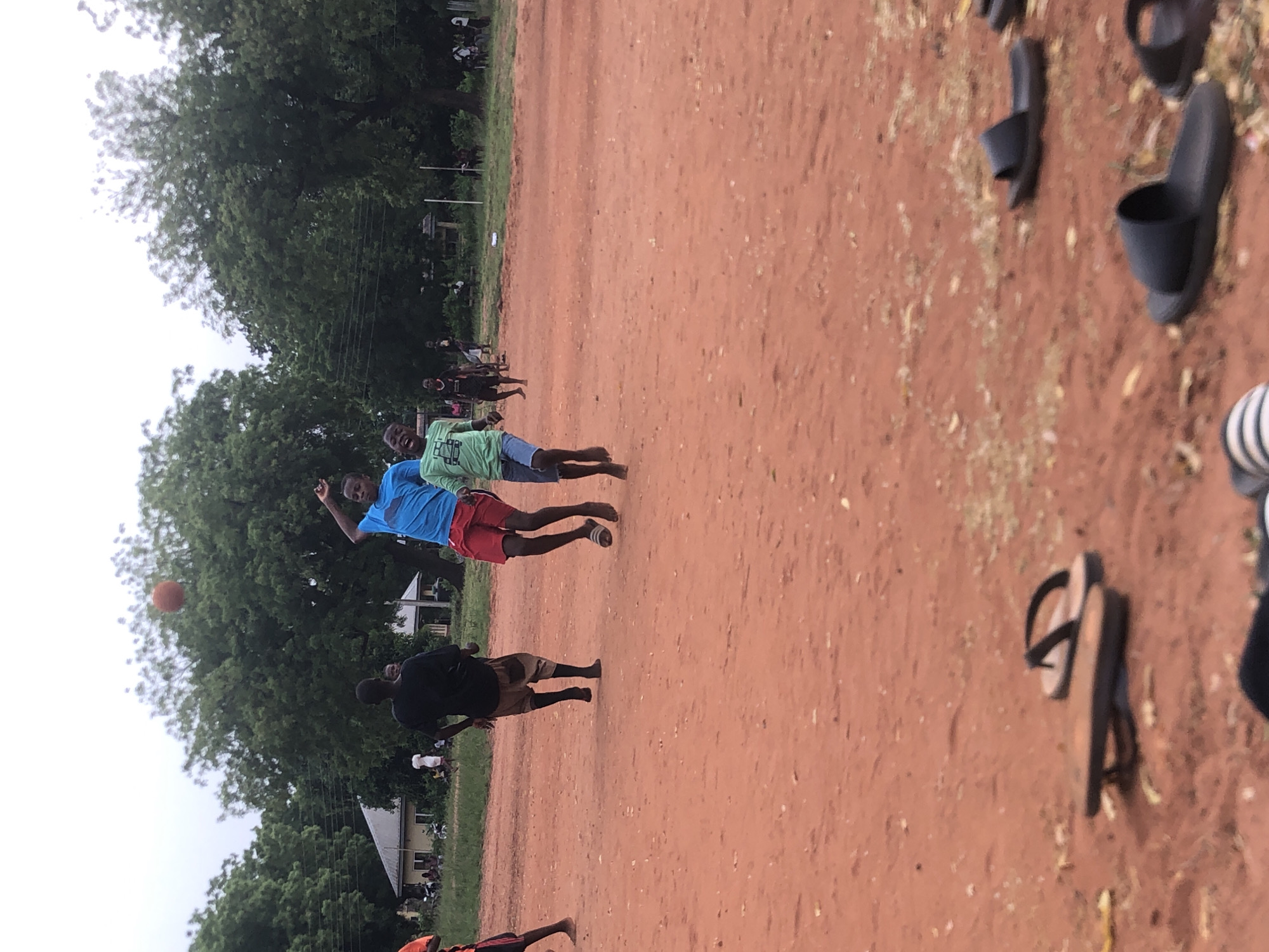 The image depicts an informal outdoor ball game taking place on a reddish-brown dirt field in Nsugbe, Nigeria. Several young males are actively engaged in play.
In the mid-ground, a boy dressed in a blue t-shirt and red shorts is captured mid-air, having jumped to reach for an orange-brown ball that is visible above his head. Standing near him is another boy in a light green t-shirt and blue shorts, observing the game. Closer to the foreground, a third boy in a black t-shirt and light brown shorts stands watching the action unfold.
The ground is composed of reddish-brown earth, characteristic of many unpaved areas. Along the right side of the frame, various black and brown sandals and slippers are scattered, suggesting players have removed their footwear to play. The background is lined with dense green trees, and glimpses of modest buildings with light walls and dark roofs are visible through the foliage.
The sky is bright and appears overcast or diffusing sunlight, indicating it is daytime. The scene conveys a lively, community atmosphere with children or young adults engaged in recreational activities in an open, natural setting. No discernible text is clearly visible on clothing or in the environment.