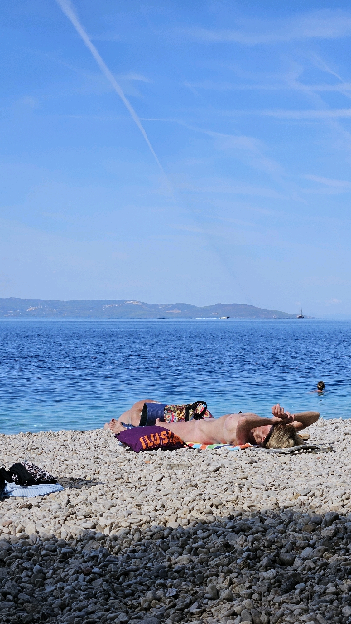A person is sunbathing on a pebbly beach in Općina Podgora, Croatia. The individual is lying on their back on a towel, with their arms raised above their head. A purple pillow with the word "ILUSA" in orange letters is placed near their feet. A patterned bag and a dark blue mat are also on the towel. In the background, the deep blue Adriatic Sea stretches towards a distant, hazy coastline with hills. The sky is a clear blue with a faint white contrail. The foreground is dominated by the texture of the pebbles. Another person is partially visible in the water in the distance. Two boats are also visible on the sea.
