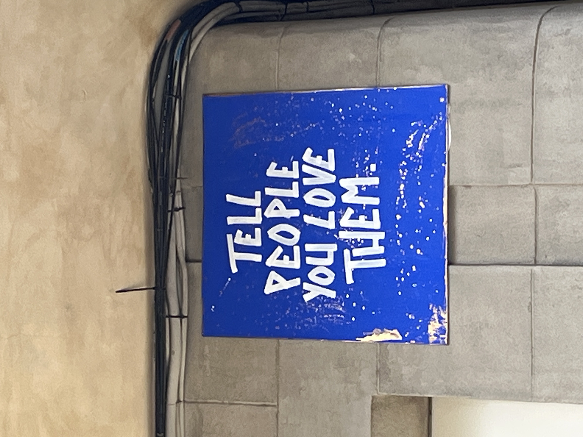 A slightly rotated close-up shows a blue sign with white lettering that says, "TELL PEOPLE YOU LOVE THEM" mounted on a gray tile wall. The sign is rectangular and positioned vertically. The background wall is constructed of large gray tiles with a textured surface. To the left of the sign is a section of a beige wall with visible textures, alongside several black and white electrical wires bundled together and secured with black zip ties. The scene suggests an indoor setting, possibly a commercial or public space. The location is Barcelona, Spain.