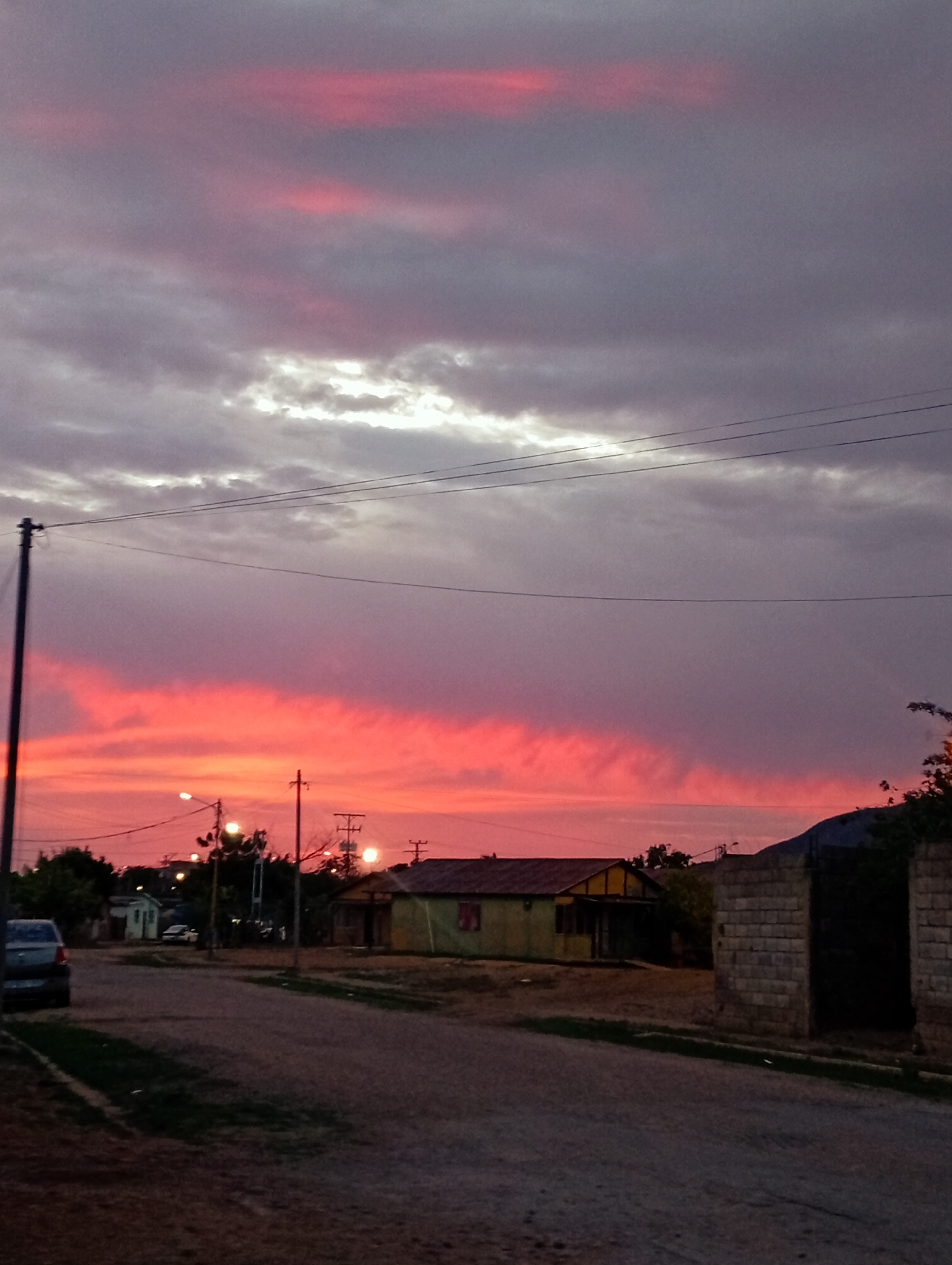 The image depicts a street scene in Juan Griego, Venezuela, during sunset. The sky is filled with dramatic clouds, with vibrant pink and red hues dominating the lower half, transitioning to muted purple and gray tones higher up. A few horizontal power lines are visible across the sky. On the left side of the street, a dark-colored car is parked. Further down the street, streetlights are illuminated, casting a warm glow. Several modest houses and structures line the street, some with visible windows and doors. A prominent brick wall with a dark opening or gate stands on the right side of the road. Vegetation, including trees, can be seen interspersed throughout the scene, particularly in the background. The overall impression is of a quiet, residential area at dusk.