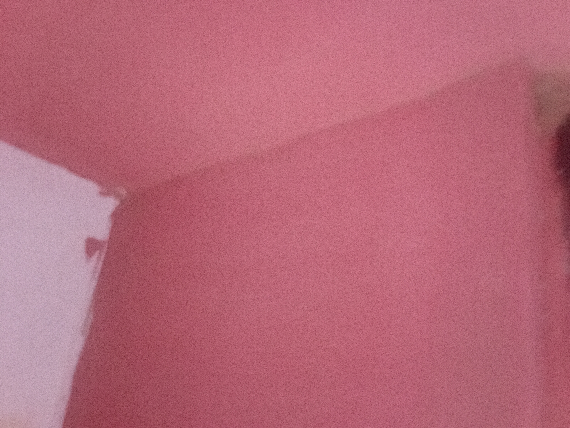The image displays a close-up, blurry view of two pink walls meeting at an inside corner. The lighting suggests an indoor environment. There is no indication of the city of Jauro Yinu, Nigeria, or any specific location within it. The scene is devoid of people, subjects, objects, actions, or interactions.