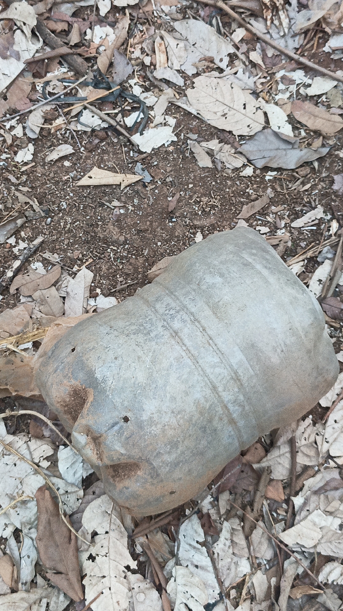 An aerial shot shows a damaged, light grey plastic container lying on its side in a dirt area covered with dry leaves and twigs. The container appears flattened and misshapen, with several horizontal ridges visible along its curved surface. A dark opening or indentation is present on the left side of the container, resembling a face or mouth, with two small holes above it. The background consists of dry soil, scattered brown and white leaves, and thin branches. The scene appears to be outdoors, possibly in a natural or undeveloped area within the city of Kandangmas, Indonesia.