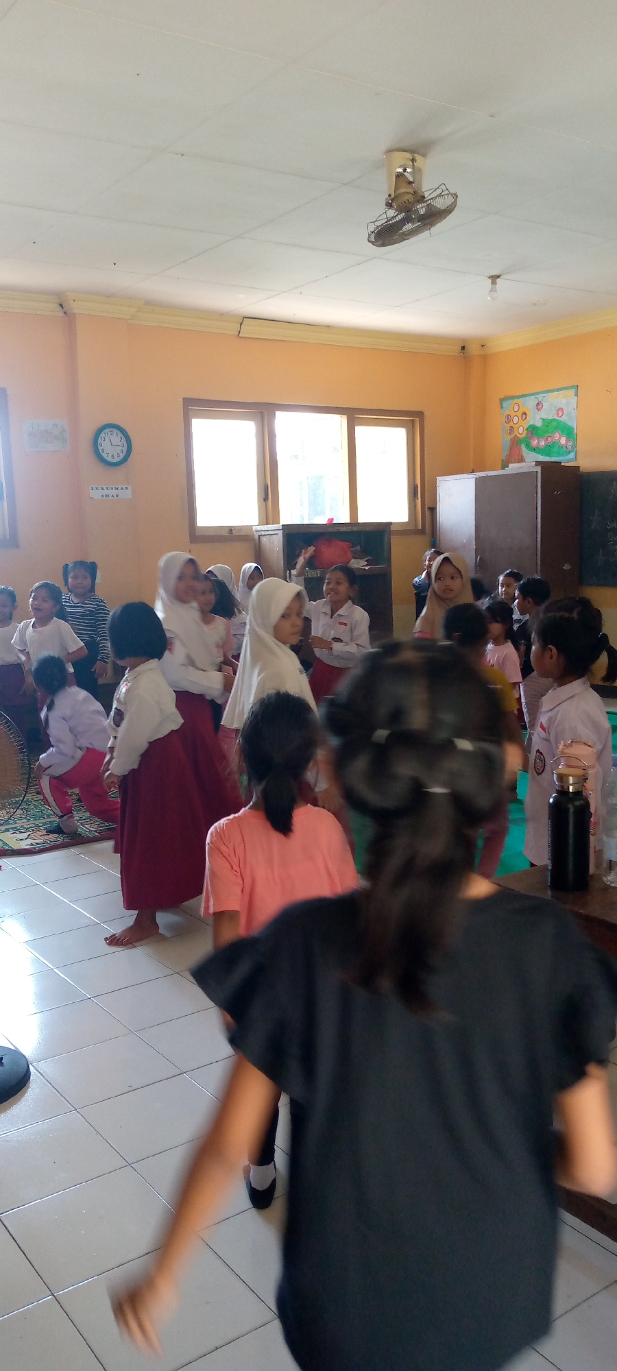 In a classroom in Pacing, Indonesia, a group of young students, mostly girls, are actively engaged in what appears to be a lively activity. The room is painted a warm, light orange and has large windows that let in natural light. The floor is tiled with white squares. A ceiling fan is visible, and a circular clock hangs on the wall, displaying the time. The students are wearing school uniforms, some with white shirts and red skirts or pants, and others with hijabs. One student in the foreground is facing away from the camera, wearing a black t-shirt. Another student in a pink t-shirt is also visible. The students are interacting and moving, suggesting a classroom lesson or playtime. A cabinet and a bulletin board with colorful drawings are also part of the classroom's furnishings. A black thermos is placed on a table on the right side of the image.