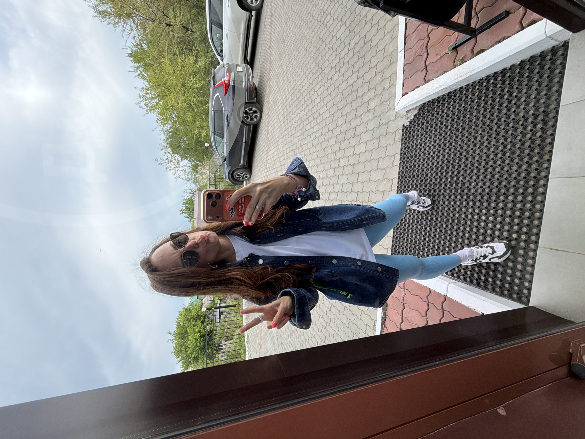 A woman is taking a selfie outdoors. She is wearing sunglasses, a denim jacket, a white t-shirt, and light blue leggings. She is holding up two fingers in a peace sign. A car is parked on a paved street in the background. The location is Birobidzhan, Russia.
