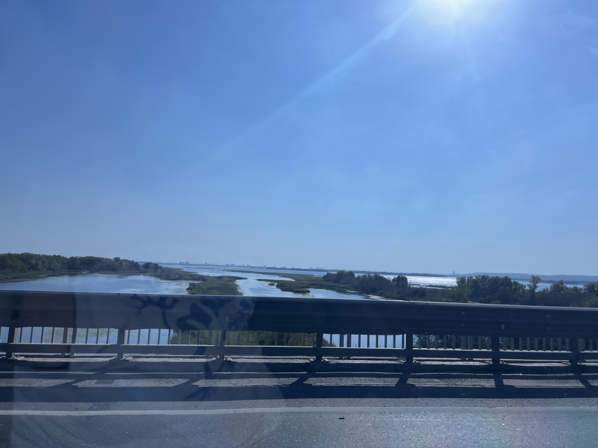 The image shows a view from a bridge overlooking a wide, calm river with islands of reeds and vegetation. The sky is clear and blue with the sun prominently visible in the upper right corner, casting a bright glare. Trees line the far bank of the river, and a city skyline is faintly visible in the distance across the water. The bridge itself has a metal railing with vertical supports, and a road surface is visible in the foreground. The image appears to have been taken on a sunny day. The location is the city of Shumeika, Russia, specifically the bridge over the Volga River.