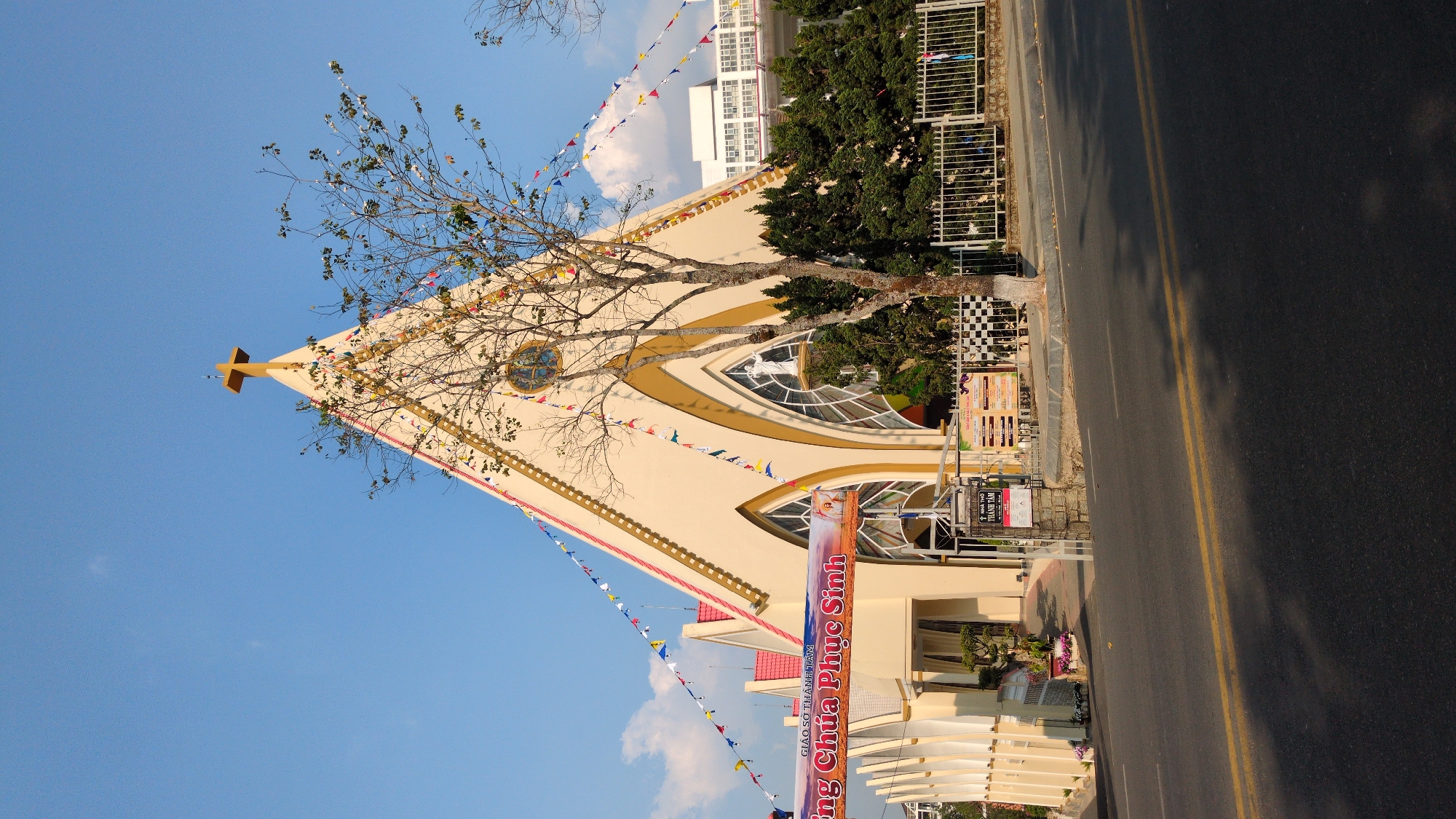 The image captures a church in Da Lat, Vietnam. The church has a pointed roof topped with a cross, a light beige color, and triangular windows with arched frames. Banners with Vietnamese writing and strings of colorful pennants adorn the building's facade. A deciduous tree stands to the side, its branches extending towards the church. Next to the church is a sidewalk with a metal fence and a building in the background. A street with double yellow lines runs in front of the church, and a clear blue sky is visible in the background.