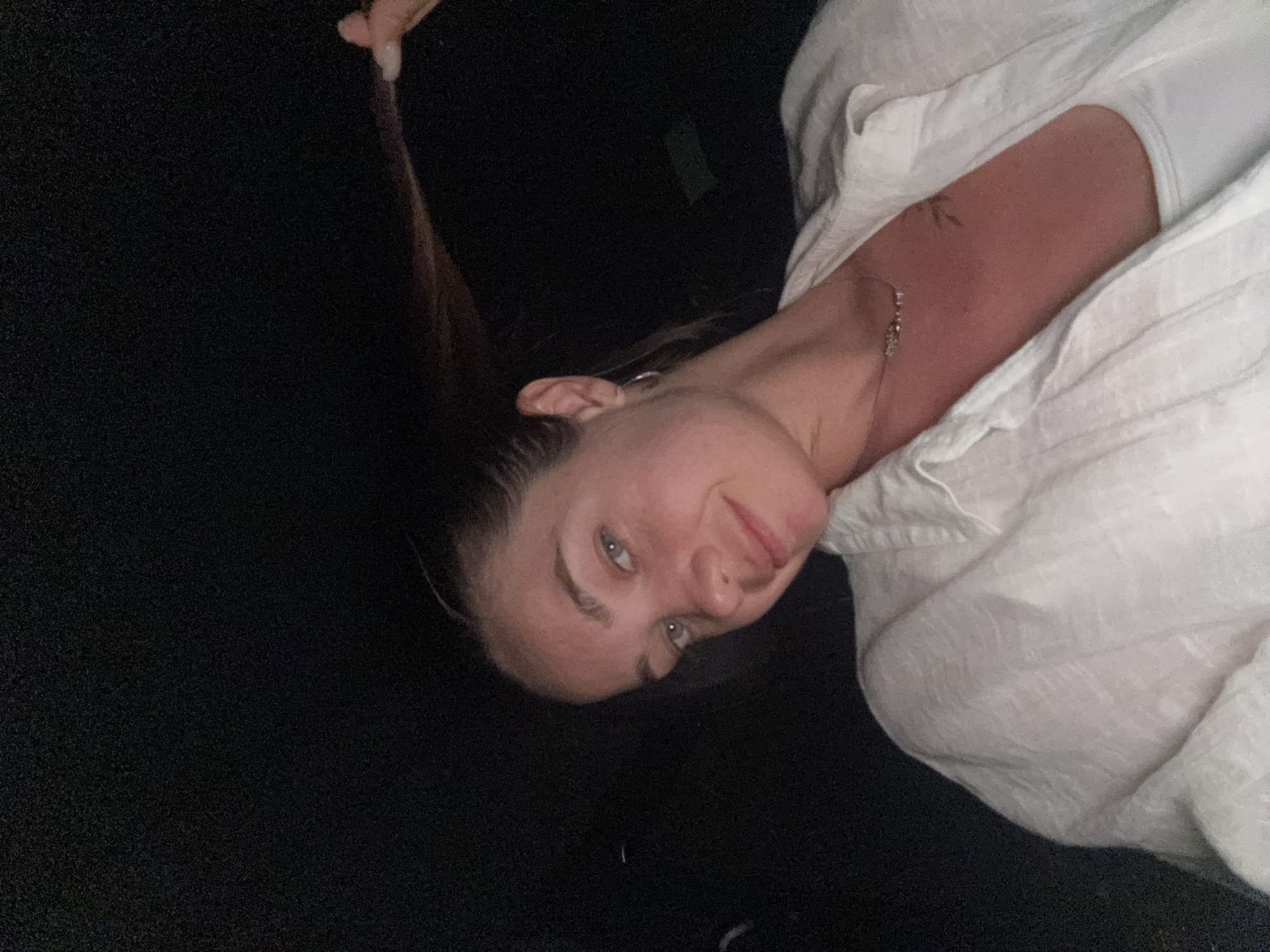 The image shows a close-up, selfie-style shot of a woman in her early twenties. She has blonde hair pulled back, with a strand in her right hand. She is wearing a white linen button-up shirt over a white tank top. A thin silver necklace with small stones is visible around her neck, and a small, dark tattoo is on her upper chest. She is also wearing a silver hoop earring in her left ear. The background is dark and out of focus, suggesting the photo was taken indoors or at night. Her skin is lightly tanned and there are no visible injuries. The location is described as Radovići, Montenegro.
