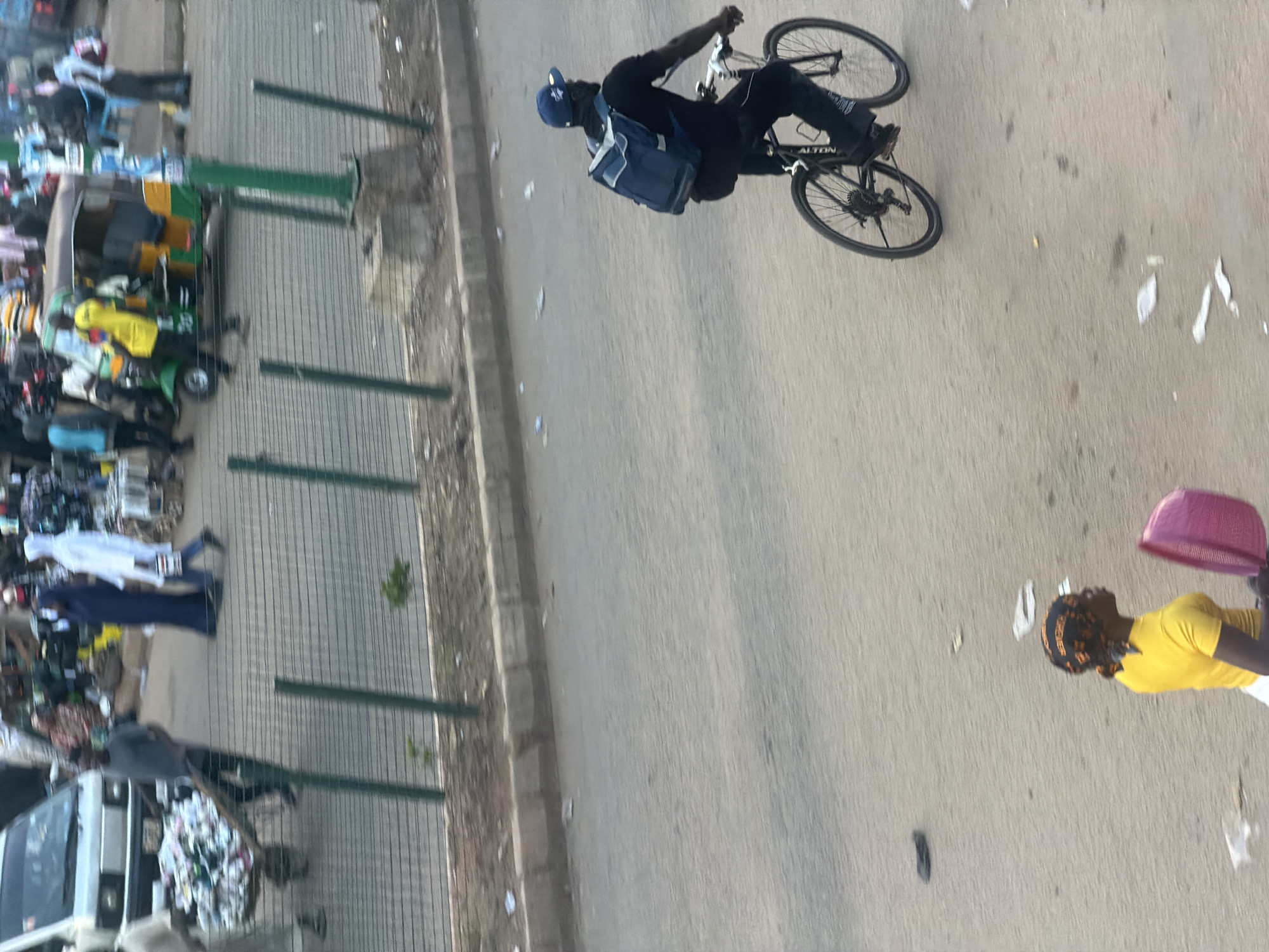 The image depicts a busy street scene in Bukuru, Nigeria, with various individuals and vehicles. In the foreground, a woman wearing a yellow shirt and headscarf is walking while holding a pink basket. Further down the street, a man on a bicycle, wearing a dark shirt and a blue baseball cap, carries a blue backpack. In the background, a white van is parked on the left side of the road, and a green and yellow auto-rickshaw is visible. Several other people, some dressed in traditional attire like a blue abaya and a white hijab, are seen walking and interacting in the area, with market stalls and various goods lining the street. The road surface is asphalt, and a metal fence runs along the side of the street. The overall impression is one of a bustling marketplace and thoroughfare.