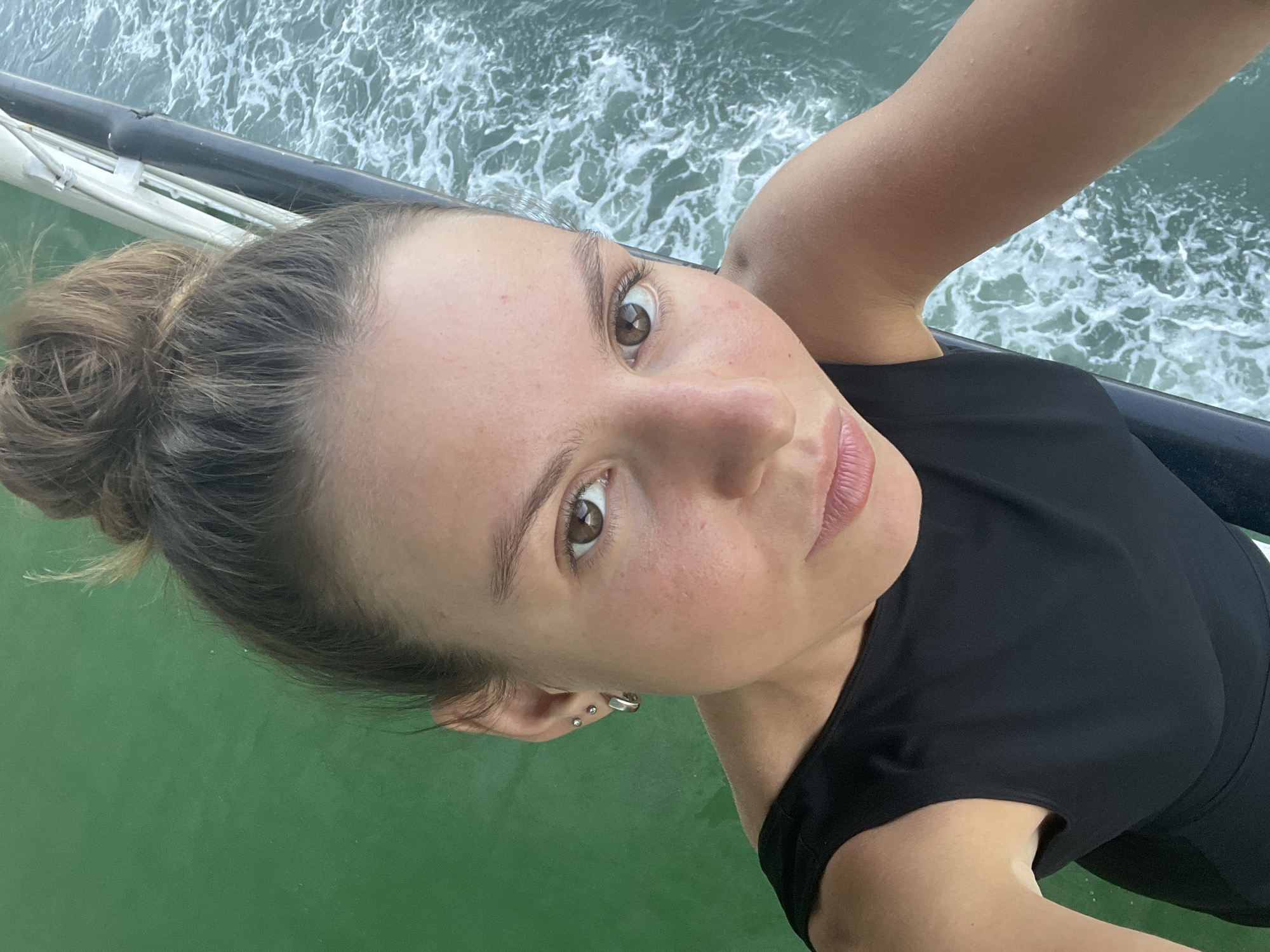 A close-up selfie captures a young woman with her hair in a messy bun, her brown eyes looking directly at the camera. She is wearing a black sleeveless top, and her right shoulder and arm are visible. She has multiple small earrings in her left ear. The background is a mix of green water on the left and turbulent blue-green water with white foam on the right, suggesting she is on a boat or ferry. A dark railing or edge of the vessel runs diagonally through the right side of the image, with white supports visible behind it. The scene appears to be outdoors, likely on the water. The image does not provide specific details to pinpoint the exact location within Batas Desa Skotong Timur, Indonesia.