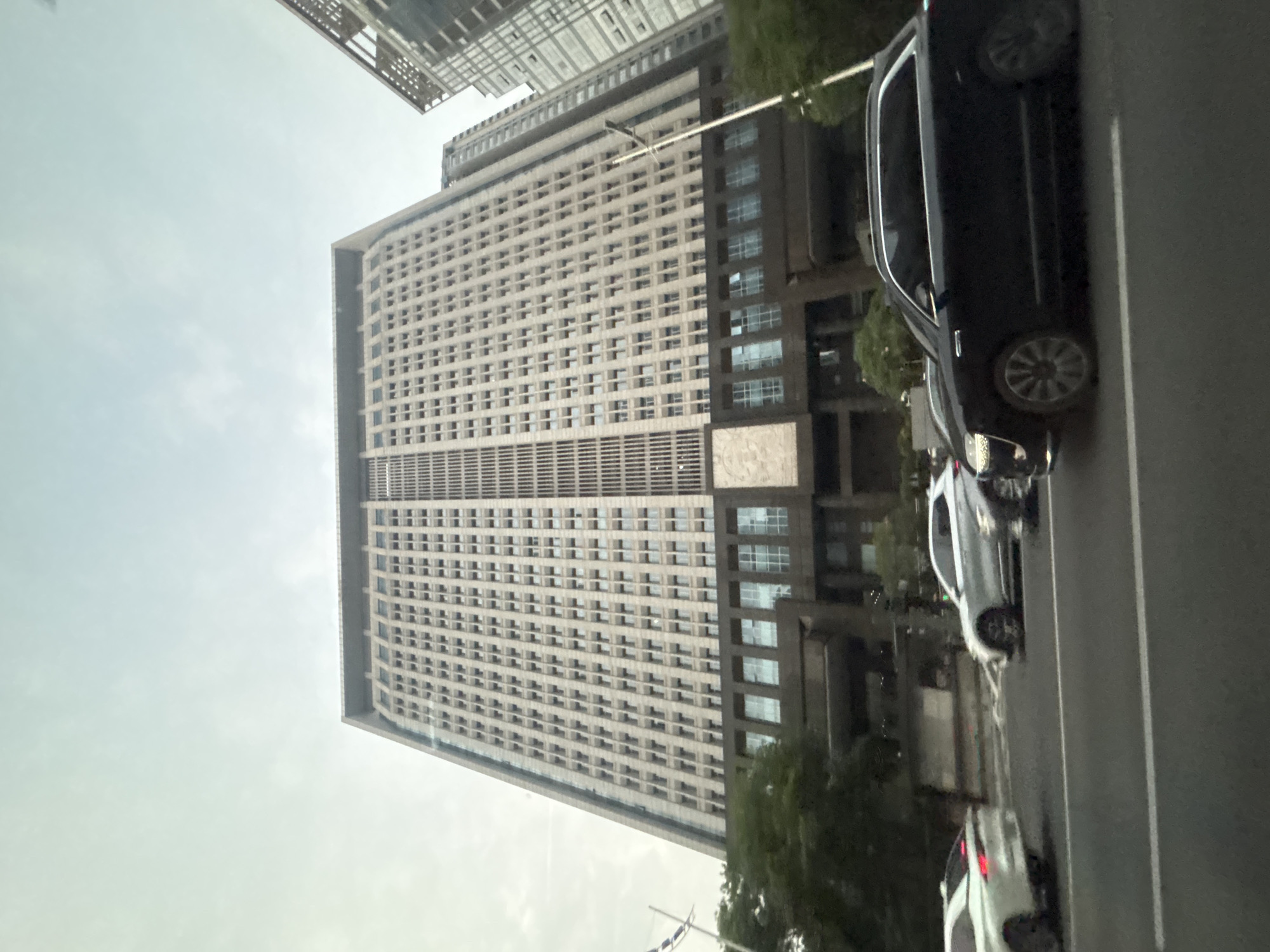 The image shows a street scene in Tianhe District, China, with a large, modern office building dominating the background. The building is a high-rise with a facade covered in numerous evenly spaced windows and vertical elements, suggesting a contemporary architectural style. In front of the building, a street is visible with several cars driving on it. The cars include a black SUV in the foreground, a white SUV to its left, and another white car further to the left, all appearing to be in motion. Trees are present on the left side of the image, providing a touch of greenery against the urban backdrop. The sky is overcast, suggesting a cloudy day. The overall scene depicts a typical urban environment with a focus on commercial architecture and vehicular traffic.