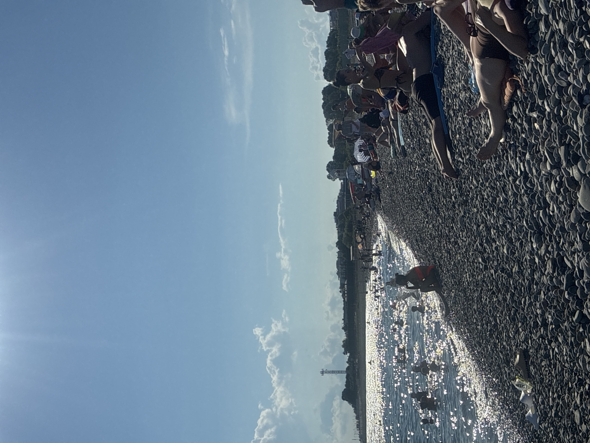 A group of people are gathered on a pebble beach, with the sea on the left and a line of trees and buildings on the right. The sun is shining brightly, casting a glittering reflection on the water, suggesting it is midday. Some people are sitting and lying on the beach, while others are in the water. A person in the water appears to be swimming or playing. The sky is clear blue with scattered white clouds. A tall structure resembling a radio tower or lighthouse is visible in the distance. The overall mood seems relaxed and leisurely, typical of a beach day. The image was taken in Sirius, Russia. There is no visible text.