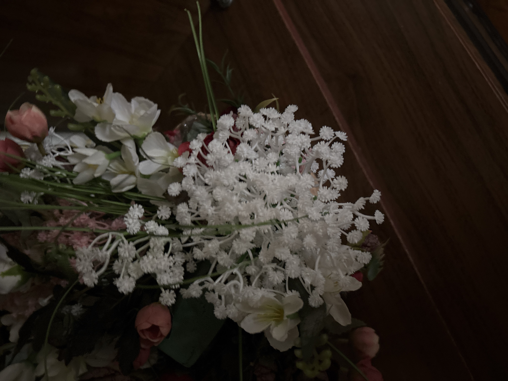 A close-up image captures a bouquet of artificial flowers set against a dark, wooden background. The central and most prominent feature is a large cluster of small, delicate white flowers, similar to baby's breath, which appear to be made of plastic or fabric and are arranged densely. Interspersed within the bouquet are several larger white flowers, possibly lilies or daffodils, with distinct petals and visible yellow stamens in their centers.
Faintly visible among the white blooms and green foliage are several light pink or peach-colored flower buds and partially open blossoms, resembling roses, adding subtle color accents. Green stems and leaves are woven throughout the arrangement, providing a naturalistic base. The lighting is dim, creating soft shadows and highlighting the texture of the white flowers, while the background recedes into darkness.
The background is a dark brown wooden surface, possibly a wall or furniture, with subtle grain lines visible, angled diagonally from the upper right to the lower left of the frame. No activity or event is taking place, as the image is a still life of a decorative arrangement. There are no people, visible text, or specific architectural or cultural cues within the image to indicate the location as Vagharshapat, Armenia.