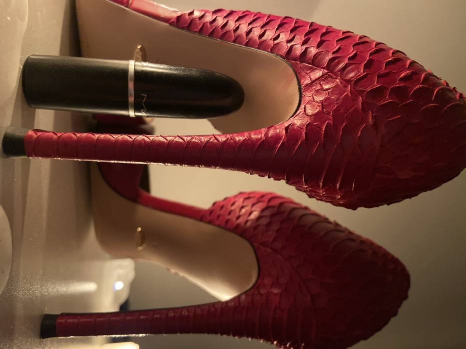 Two high-heeled, red, snakeskin shoes are stacked. The first shoe is upside down with the heel facing up and the toe facing down. On top of the heel is a black tube of lipstick with silver accents. The second shoe is beneath the first and oriented correctly, toe upward and heel downward. The heels are narrow and red with black tips, and the interior of the shoes is tan.