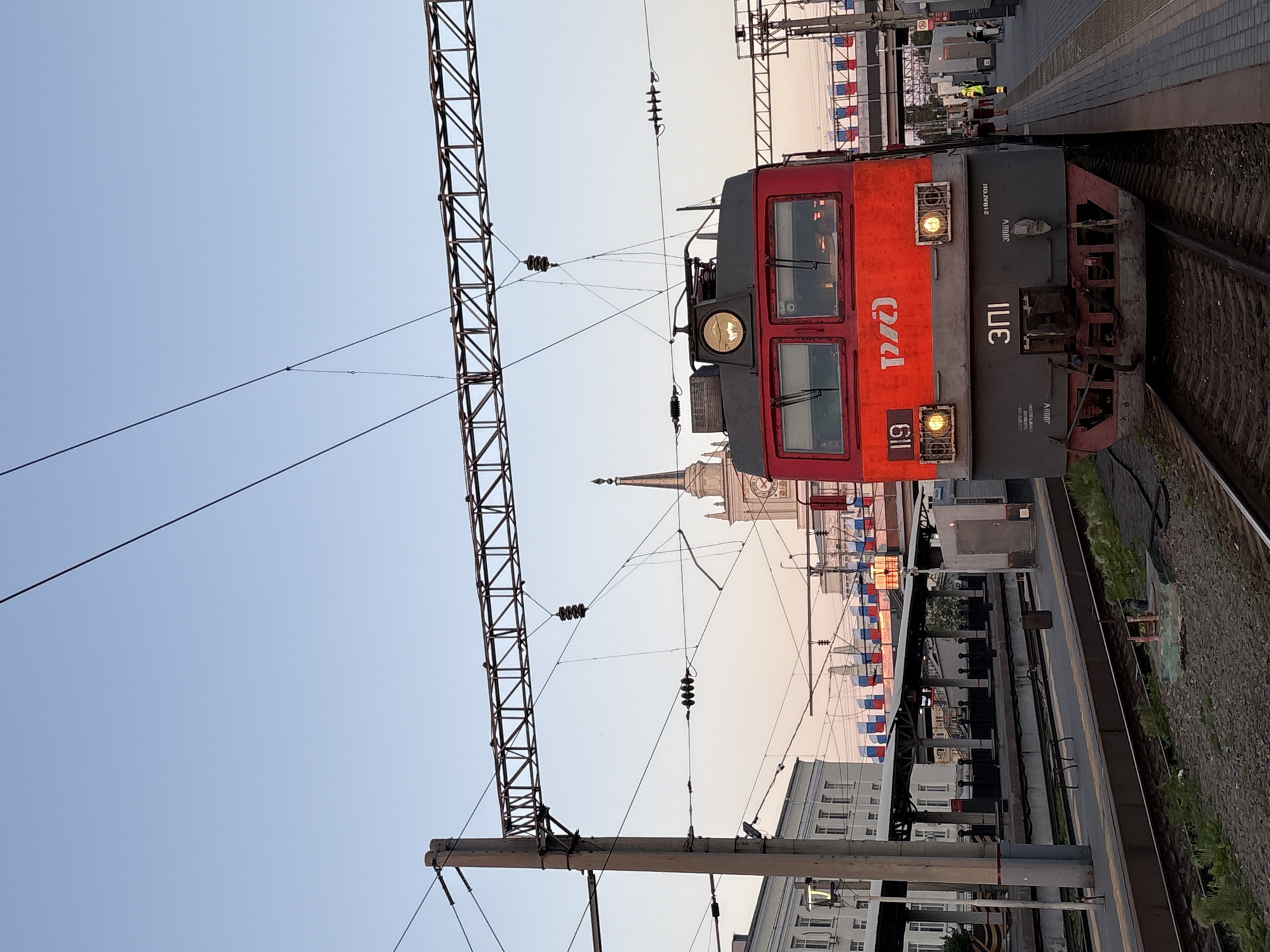 A red and gray electric locomotive, labeled "РЖД" (Russian Railways) and "119" on the front, is positioned at a train station. The locomotive has two headlights illuminated and a series of Cyrillic characters and numbers on its lower front panel. In the background, a tall clock tower is visible, likely part of a building. The station platform is paved with gray and reddish-brown tiles, with tracks running in the foreground. Overhead power lines crisscross the scene. Along the platform and in the distance, strings of Russian flags are displayed. The sky is a clear, pale blue, transitioning to a pale pinkish hue near the horizon, suggesting it is either early morning or late evening. A few individuals, indistinct in the background, are present on the platform. The location is identified as Volgograd-1 station in Volgograd, Russia.