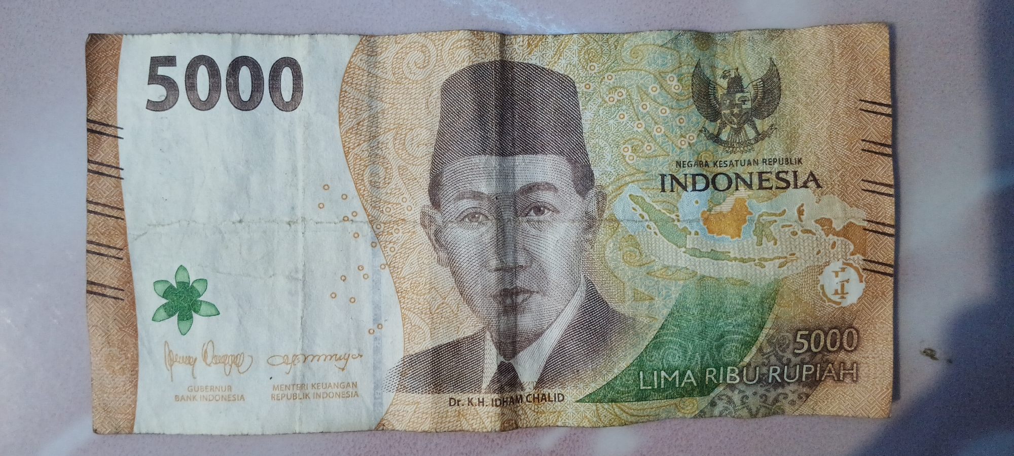 The image shows a folded Indonesian 5000 Rupiah banknote. The banknote features a portrait of Dr. K.H. Idham Chalid. Text on the banknote includes "BANK INDONESIA", "GUBERNUR", "MENTERI KEUANGAN", "REPUBLIK INDONESIA", "LIMA RIBU RUPIAH", and "INDONESIA". The national emblem of Indonesia, Garuda Pancasila, is also visible. The banknote is placed on a surface that appears to be a table or floor. There is no information in the image that indicates the location is the city of Rejing, Indonesia.