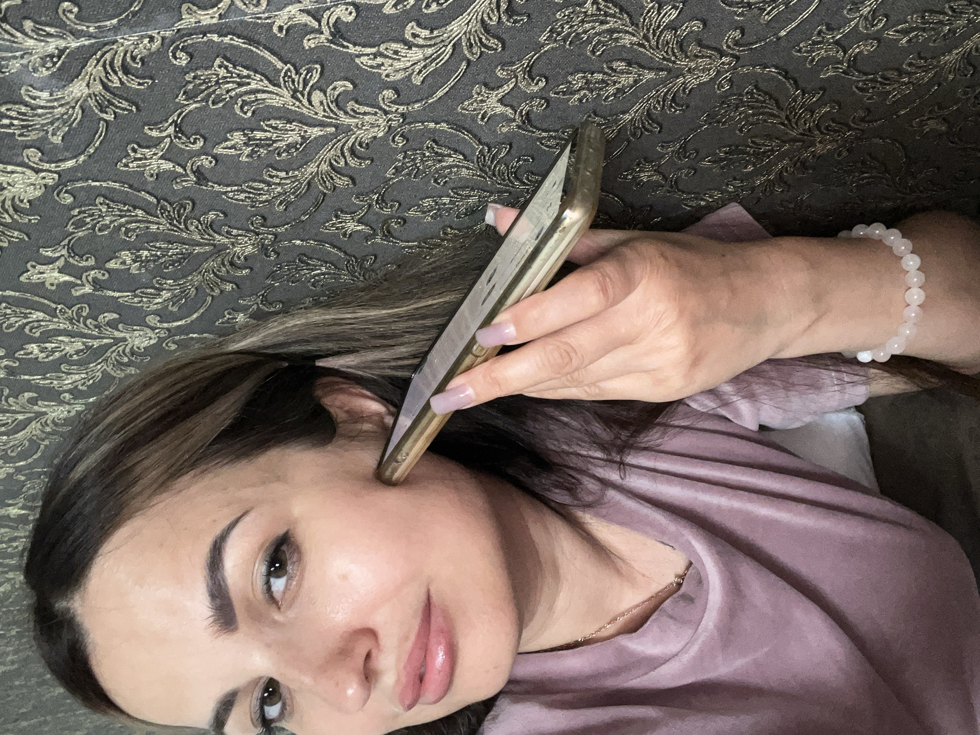 A medium shot shows a woman with dark hair and light skin holding a gold-colored smartphone to her right cheek. Her right hand is positioned to support the phone, with her fingers wrapped around its edge and her thumb resting on the screen. She is wearing a purple t-shirt and a white beaded bracelet on her left wrist. Her left hand is not visible. The background is a dark wall with a gold floral pattern. The scene is indoors, with no indication of a specific city or location.