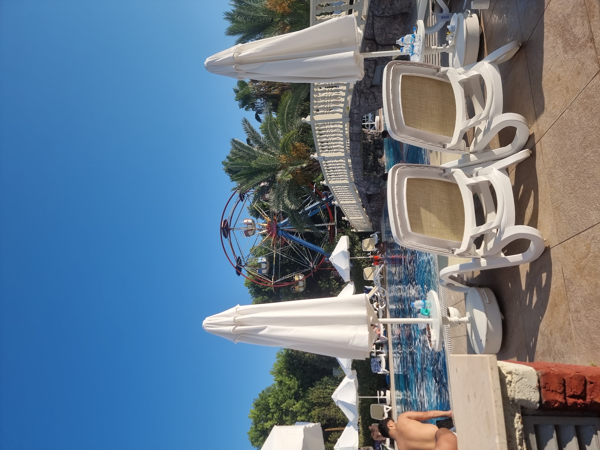 The image shows a view of a swimming pool area at what appears to be a resort in Manavgat, Turkey, with a Ferris wheel in the background. Two white lounge chairs are in the foreground, facing away from the camera. Behind them, a swimming pool is visible with several people in it. A person is standing at the edge of the pool on the left side of the frame, facing away from the camera. Several white beach umbrellas are also scattered around the pool area. In the background, a colorful Ferris wheel is prominently displayed, surrounded by palm trees and lush greenery. A stone bridge with white railings crosses over a section of the pool. The sky is clear and blue.