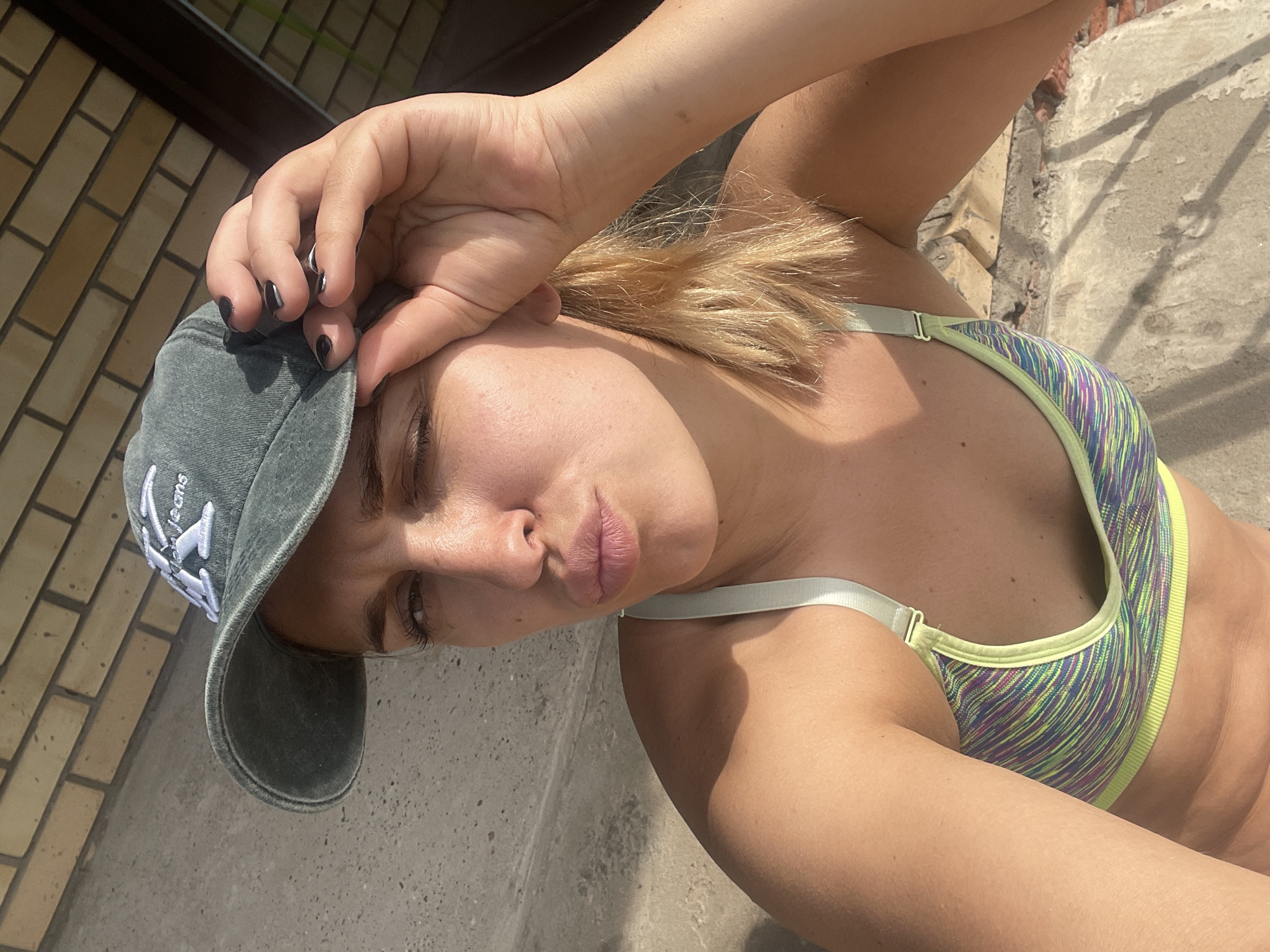 A fair-skinned woman with blonde hair is pictured outdoors, possibly in the city of Chaikovsky, Russia. She is wearing a sports bra with yellow, purple, and green stripes, and a grey baseball cap with white embroidery. Her left hand is touching the brim of the cap, while her right arm is extended to the side. The background includes a brick wall on the left and a concrete surface on the right.