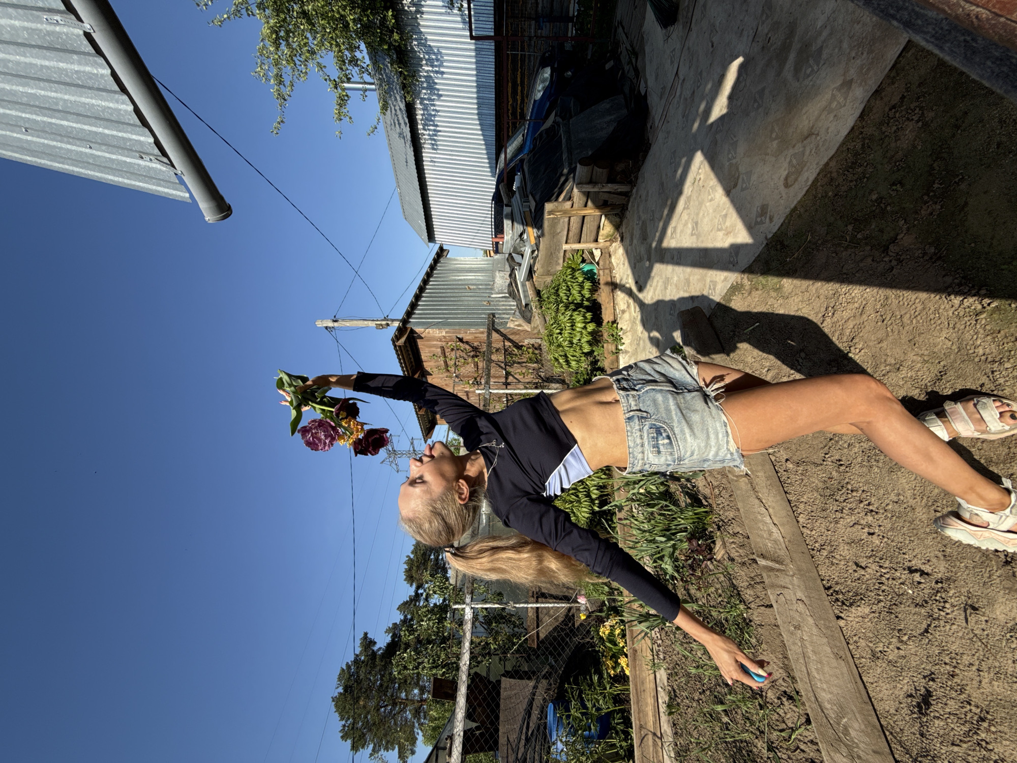 A woman in a black long-sleeve crop top and denim shorts holds a bouquet of flowers against a bright blue sky. She stands in a yard with a shed, concrete patio, and foliage. The location is in Chaikovsky, Russia.