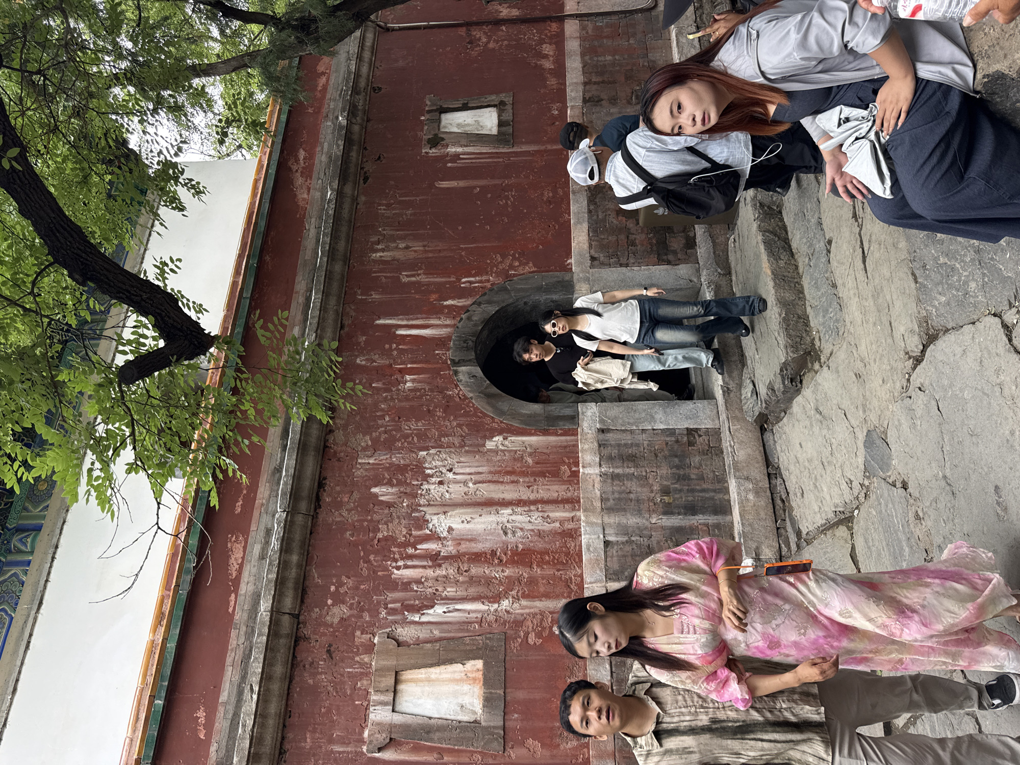 The image depicts a bustling scene at a historical site within the Haidian District of China, likely the Summer Palace (Yiheyuan), characterized by traditional Chinese architecture. A prominent, weathered red wall with peeling paint and vertical streaking forms the backdrop, featuring an arched entrance near its center. Two recessed, rectangular, off-white panels, resembling sealed windows, are embedded in the red wall, one to the left and one to the right of the archway. Above the red wall, a horizontal stone band supports a tiled roof section with green and orange tiles, topped by a white wall and a more ornate, multi-colored roof structure (partially visible) featuring blue and green patterns.
Numerous individuals of East Asian descent are present. In the immediate foreground, a man in a light-colored striped shirt and beige pants walks alongside a woman wearing a flowing, long pink and white tie-dye patterned dress. She holds an orange device, possibly a smartphone, attached to a strap. Both are moving from left to right.
Through the arched entrance, a man in a dark t-shirt and light pants is visible, followed by a woman in a white t-shirt, blue jeans, and white-framed sunglasses. Further into the archway, other indistinct figures can be seen. To the right of the archway, several people are navigating irregular stone steps. One man, seen from behind, wears a white baseball cap, a light striped shirt, and a black backpack.
In the foreground right, a woman with reddish-brown hair is seated on a stone step, looking towards the right side of the frame. She wears a light grey zip-up jacket over a dark blue or grey jumpsuit or wide-leg pants and holds a partially visible clear plastic water bottle. The ground consists of uneven stone paving and steps. Large, verdant trees with dense foliage are visible in the upper right and upper left portions of the frame, providing shade and partially obscuring the upper parts of the building and sky. A thick tree trunk is prominent in the upper right.