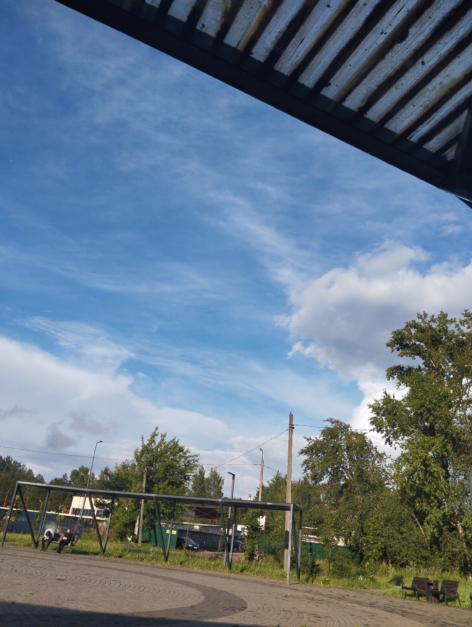 The image captures an outdoor scene in Kingisepp, Russia, under a partly cloudy blue sky. In the foreground, a paved area with a circular pattern is visible, leading to a park-like setting with green grass and trees. A metal structure, possibly a playground or a seating area, is present with two individuals sitting on what appears to be a swing set. They are dressed in casual clothing. Further in the background, there are more trees, a fence, buildings, and parked cars. A power line and utility pole are also visible. The top of the frame is partially obscured by an overhead structure with corrugated material.