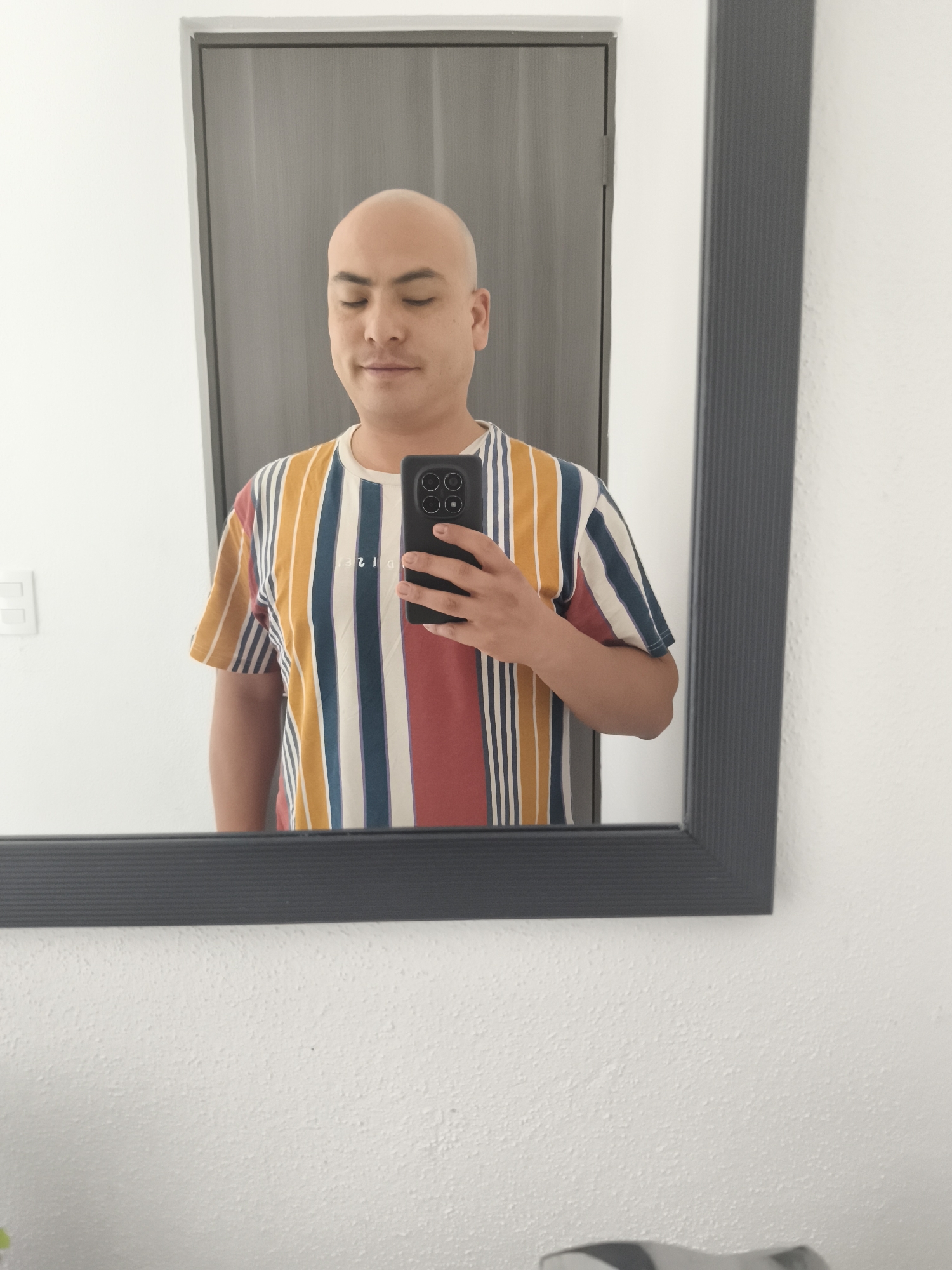 A bald-headed person is taking a selfie in a mirror. The person is wearing a striped t-shirt with a variety of colors. The mirror has a dark gray frame. The background consists of a white wall and a gray door. The location is likely inside a residence in El Carmen, Mexico.