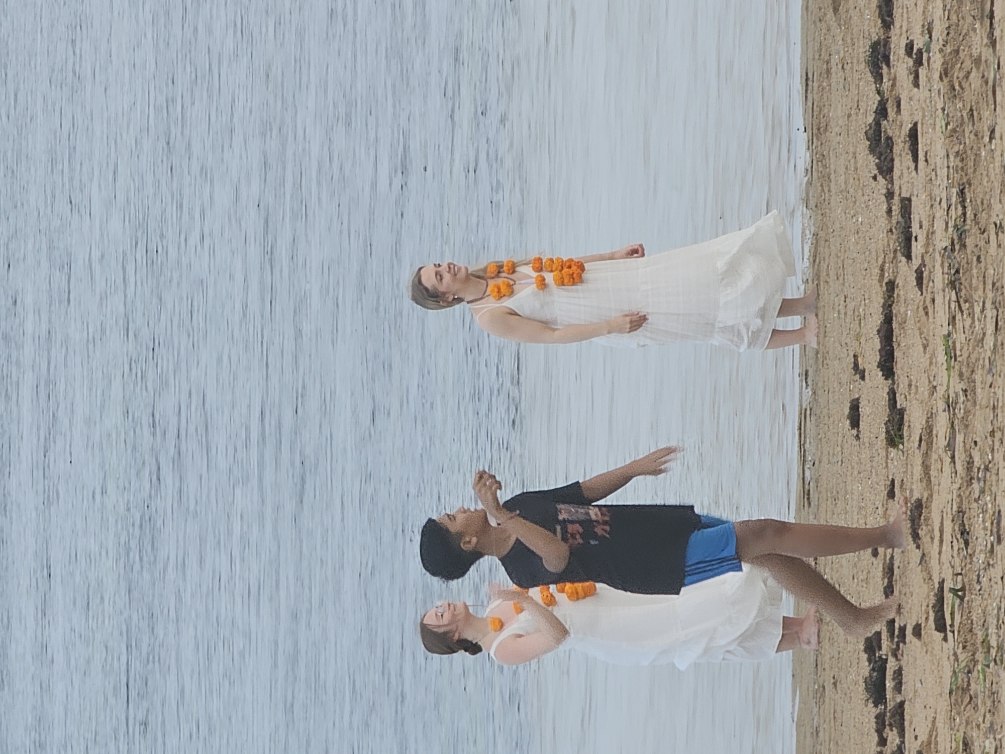 Three individuals are standing at the edge of the water on a sandy beach. The person in the foreground is a young male wearing a black t-shirt and blue shorts, facing to the right and gesturing with his right hand. To his left, a woman in a white dress is partially visible, also facing right and wearing an orange flower garland. Further to the right, another woman in a white dress, also wearing an orange flower garland, is looking to her right and smiling. The water is calm and light blue. The beach is sandy with some scattered seaweed. The overall scene appears to be a casual gathering or ceremony at the seaside. Based on the attire and flower garlands, the location may be Denpasar, Indonesia, possibly near a beach such as Seminyak Beach or Kuta Beach, where such cultural elements are common.