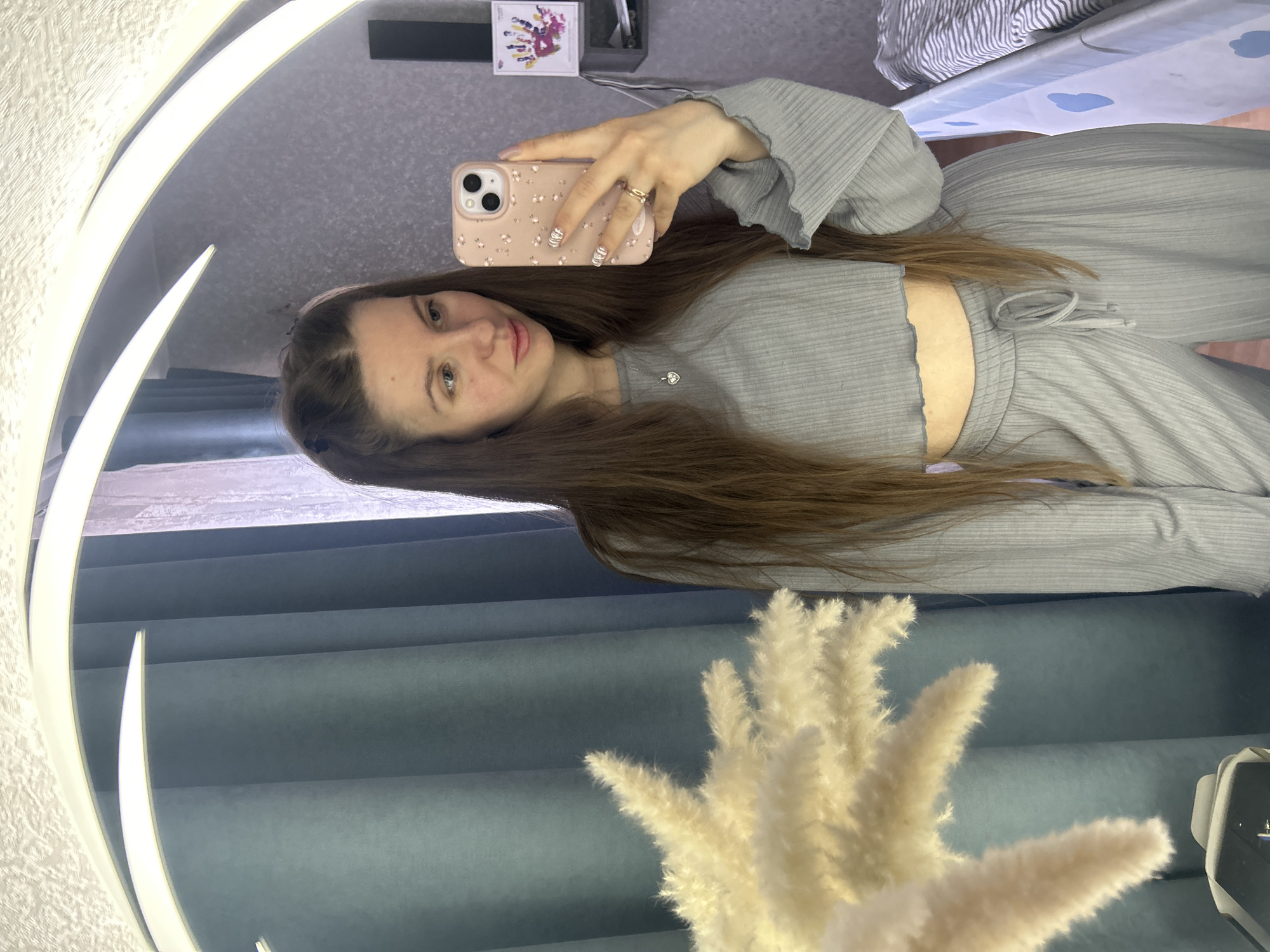 A woman is taking a selfie in a mirror. She has long brown hair and is wearing a light gray outfit. She is holding a pink cell phone with a floral pattern on the case. The background includes a light gray wall, a blue curtain, and a white decorative plant. On a shelf above the woman's head is some kind of artwork and what appears to be a cable box. In the upper right, bedding can be seen. The location is Klin, Russia.