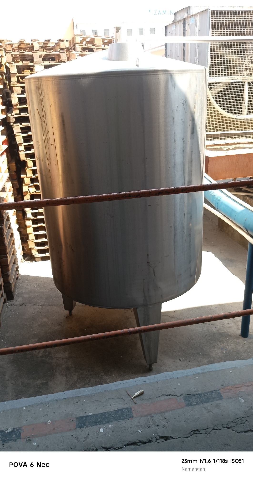 An outdoor scene in Namangan City, Uzbekistan, features a large, cylindrical stainless steel tank supported by three legs. The tank is positioned on a concrete surface, with stacks of wooden pallets to its left. A rusty metal pipe runs horizontally across the front of the tank, approximately one-third of the way up from the bottom. In the background, parts of industrial machinery, including a large fan within a caged structure, are visible. A blue pipe extends upwards to the right of the tank. The sky above is bright and appears overcast or hazy. The overall impression is an industrial or storage area, possibly outdoors.