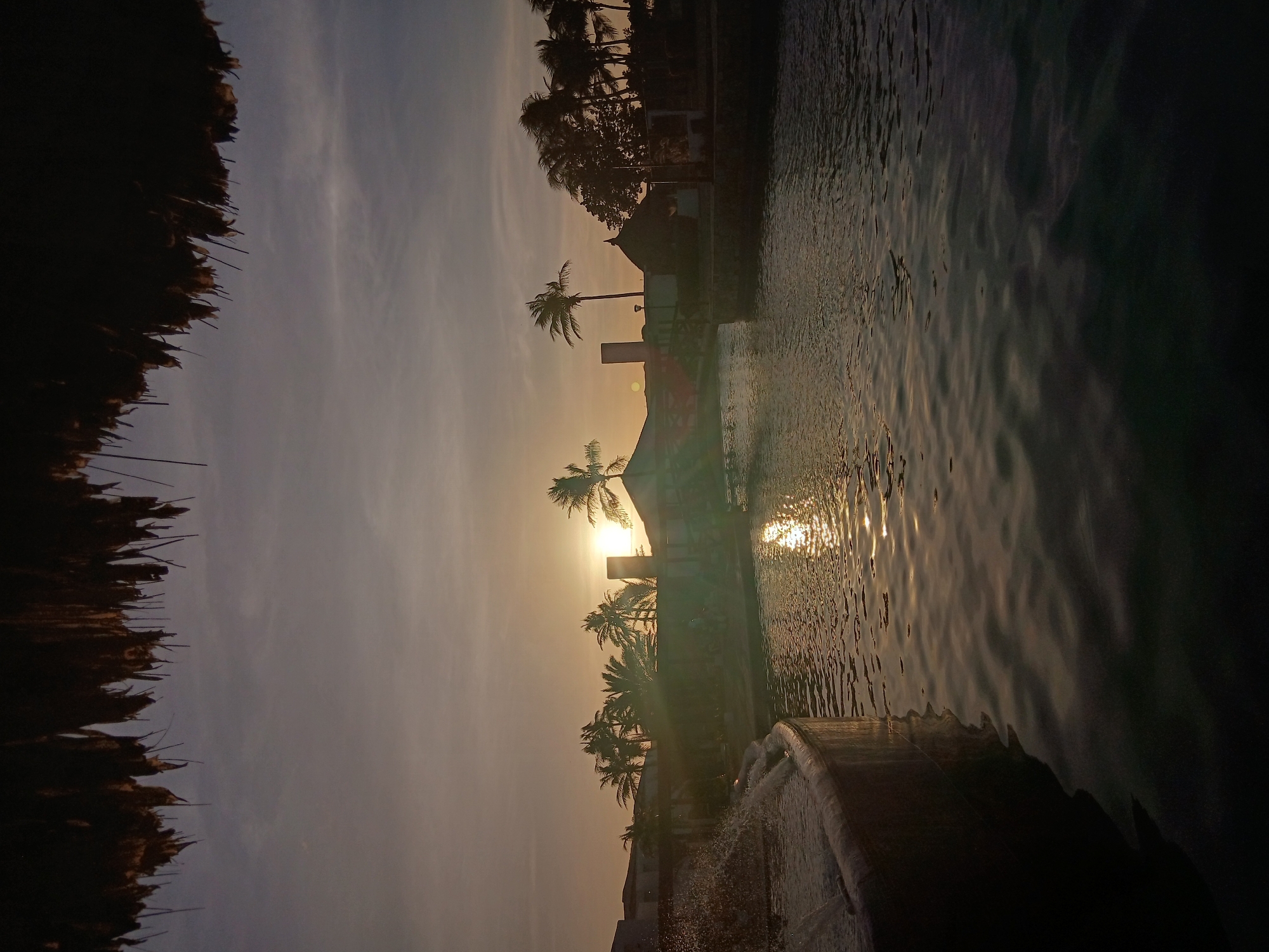 The image captures an outdoor canal or large pool environment during either sunrise or sunset, viewed from beneath the dark, silhouetted overhang of a thatched roof that spans the top of the frame. The sky transitions from a bright, warm orange-yellow near the horizon to a subdued grey-blue above, with scattered thin cloud formations. The sun, a brilliant orb positioned slightly left of center on the horizon, casts intense glare and a pronounced, shimmering reflection path across the rippled water surface.
In the left foreground, a curved, concrete-edged structure functions as a fountain, with a stream of water actively splashing upwards and outwards, illuminated by the low sun. Across the body of water, the background is dominated by a collection of structures and dense tropical vegetation, largely appearing as dark silhouettes against the bright sky. Visible structures include a building on the left with a partially obscured facade, a tall, slender, dark rectangular column, and a smaller hut or building to the right featuring a dark, conical or pyramidal thatched roof. Numerous palm trees are prominent throughout the background, their dark fronds delineated against the bright sky. Additional dark, dense foliage is present. A dark, low-lying structure, possibly a bridge or walkway, spans a section of the water in the mid-right. No human figures are present or discernible within the scene. The architecture, canal system, and tropical flora suggest a resort or residential development within the city of Altagracia, Venezuela, likely a planned community with waterways, consistent with areas such as Pueblo Viejo.