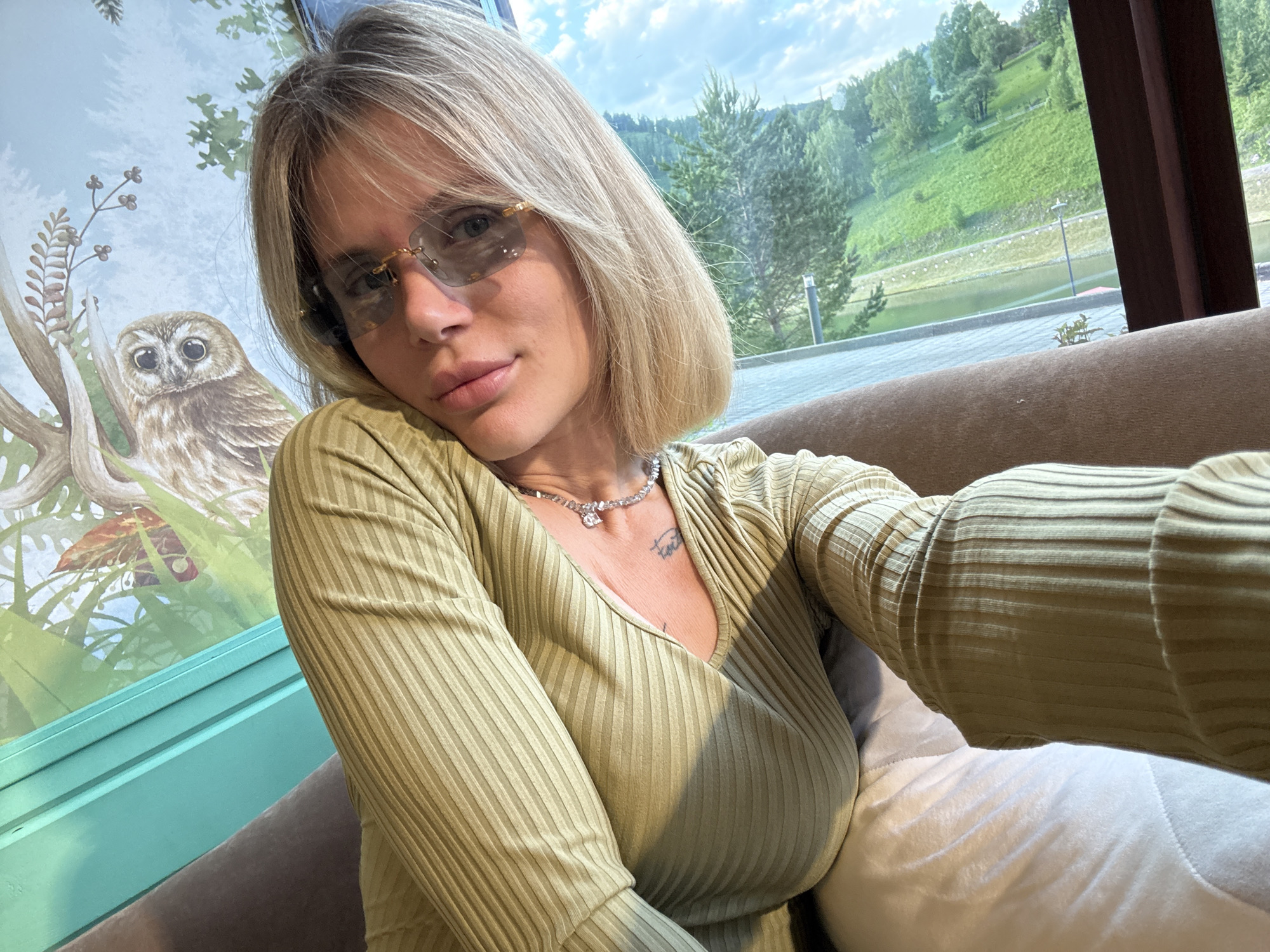 A woman with shoulder-length blonde hair and rimless, tinted sunglasses poses for a selfie while seated on a brown sofa indoors. She is wearing a light olive-green ribbed long-sleeved V-neck top and a silver-toned necklace with a small pendant. A small, illegible tattoo is visible on her upper chest.
To her left, a wall features a decorative mural depicting a wide-eyed, small brown owl perched on a branch amidst green foliage and berry-like clusters, set against a light blue background. To her right, a large window reveals a scenic outdoor landscape. Beyond the window, rolling green hills covered with abundant pine trees and other vegetation extend under a partially cloudy blue sky. At the base of the hills, a body of water, possibly a lake or river, is visible, bordered by what appears to be a paved area or road. The overall setting combines a cozy indoor space with a vibrant natural view, consistent with a location like Urlu-Aspak, Russia, known for its scenic landscapes. The time of day appears to be bright daylight. Her expression is neutral and relaxed.