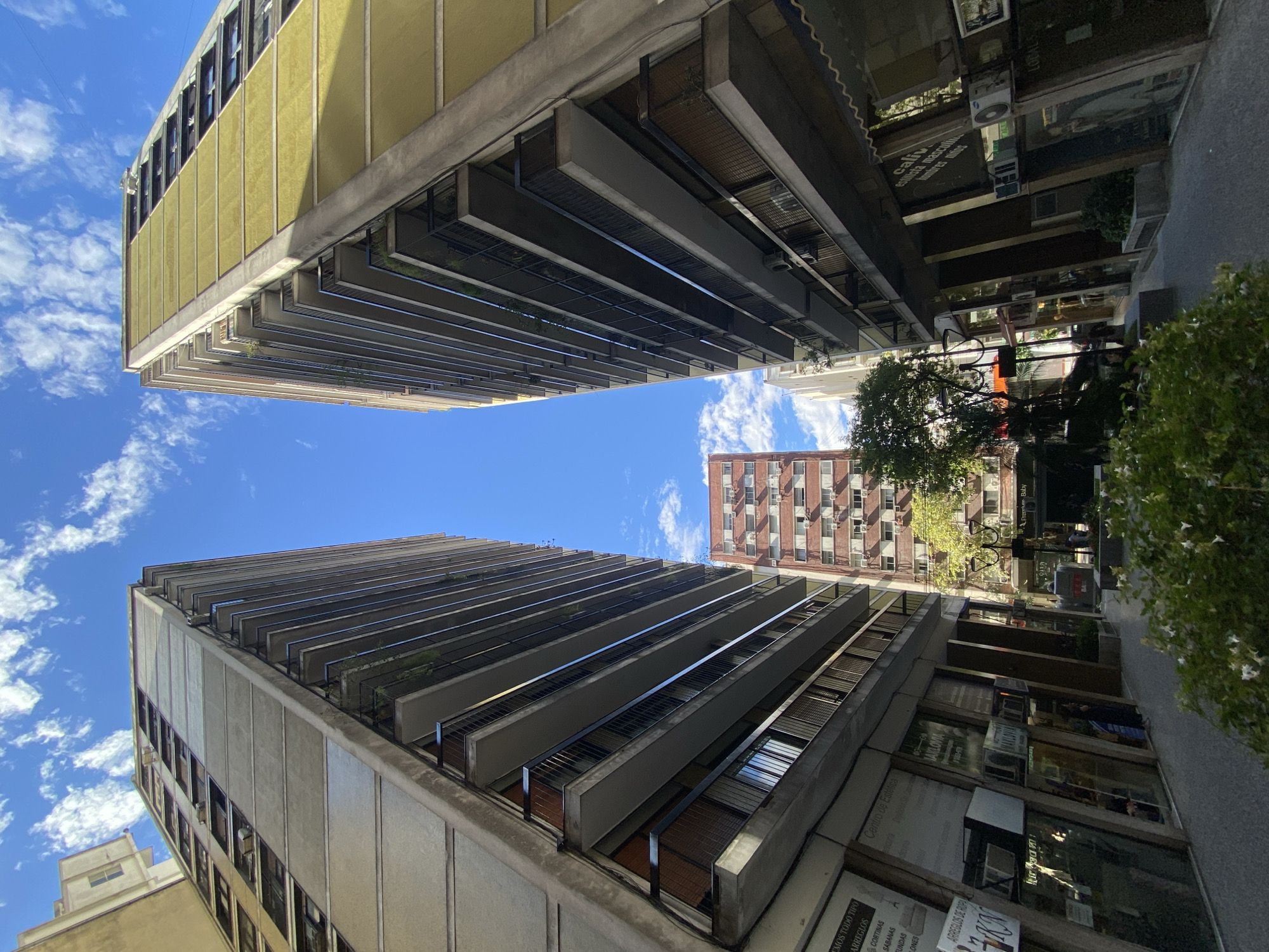 The image depicts a low-angle, upward view of a narrow urban thoroughfare in Buenos Aires, Argentina, likely situated in the Microcentro area, possibly near Florida Street or Tucumán Street. Two multi-story buildings, characteristic of 20th-century modernist architecture, dominate the foreground, symmetrically framing the sky and a distant building.
The building on the left exhibits a concrete facade in light grey tones, featuring numerous balconies with black metal railings extending along its height. Its ground floor is occupied by commercial storefronts with distinctive slanted glass panels and visible signage, including "ARREGLOS DE ROPA" (clothing alterations).
The building on the right mirrors the structural design but features a yellowish-green facade. It also presents multiple balconies with black railings and ground-floor commercial units with slanted glass windows, displaying signs such as "Centro de Estética" and "NAYLOVIA." Air conditioning units are visible on the exterior of both lower buildings.
In the background, positioned between the two foreground structures, a taller, reddish-brown building with multiple windows and numerous external air conditioning units is visible. The sky overhead is clear blue with scattered white cumulus clouds.
At street level in the immediate foreground, green foliage with small white flowers is present, consistent with a planter or small garden. Further down the street, a small tree with green leaves is visible. Indistinct human figures can be seen through the glass of some commercial establishments and on the street itself, indicating activity, though specific actions or interactions are not discernible. The ground appears paved.