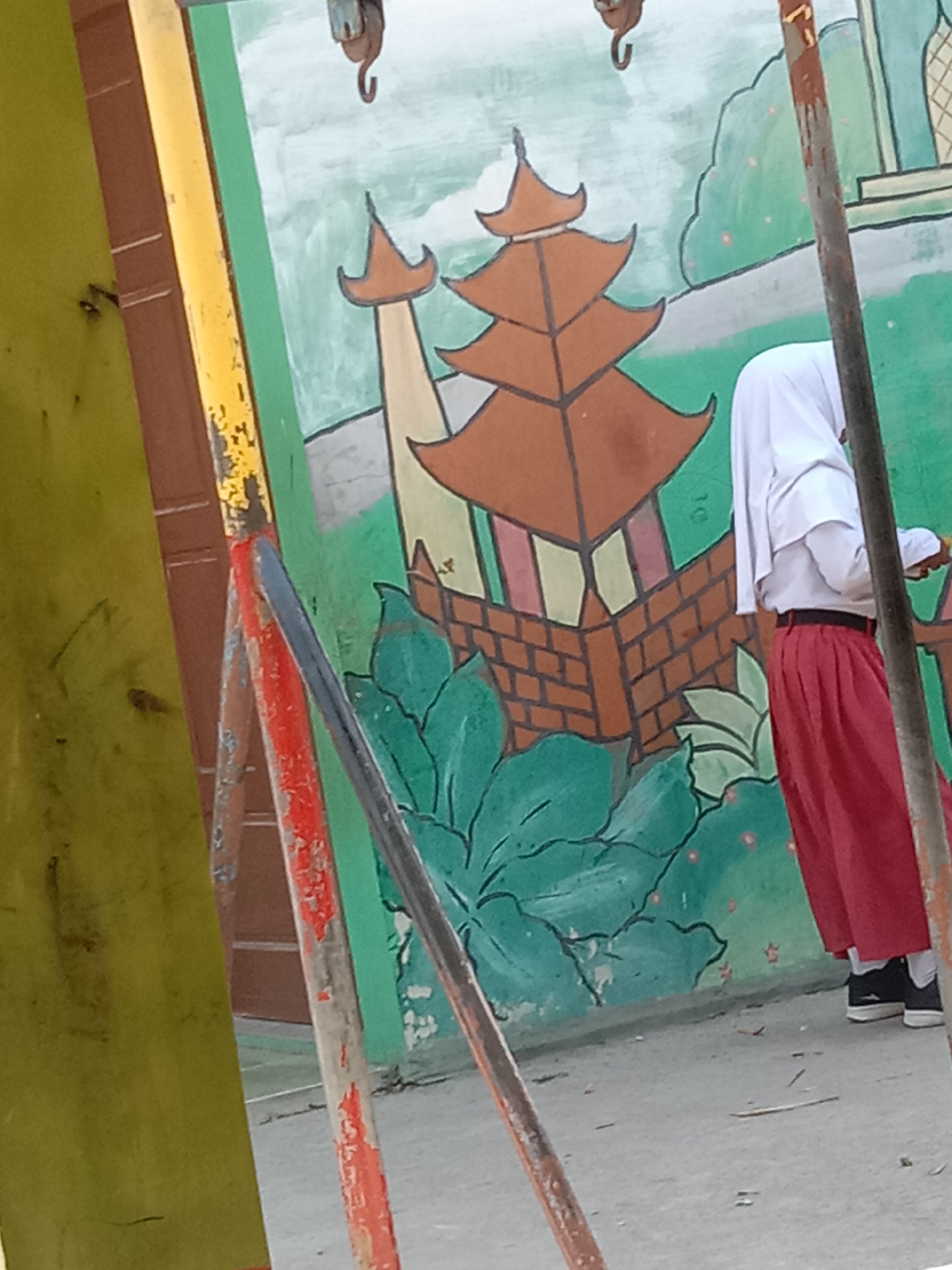 A young person, wearing a white headscarf and a red skirt, stands in front of a mural depicting a traditional building with orange tiered roofs and green foliage. The mural is painted on a wall that appears to be outdoors, with a grey concrete ground visible. Metal poles, some painted red and yellow, are positioned in the foreground, partially obscuring the mural and the person. The overall scene suggests an educational or recreational space, possibly a schoolyard, in Tanjungsari, Indonesia.