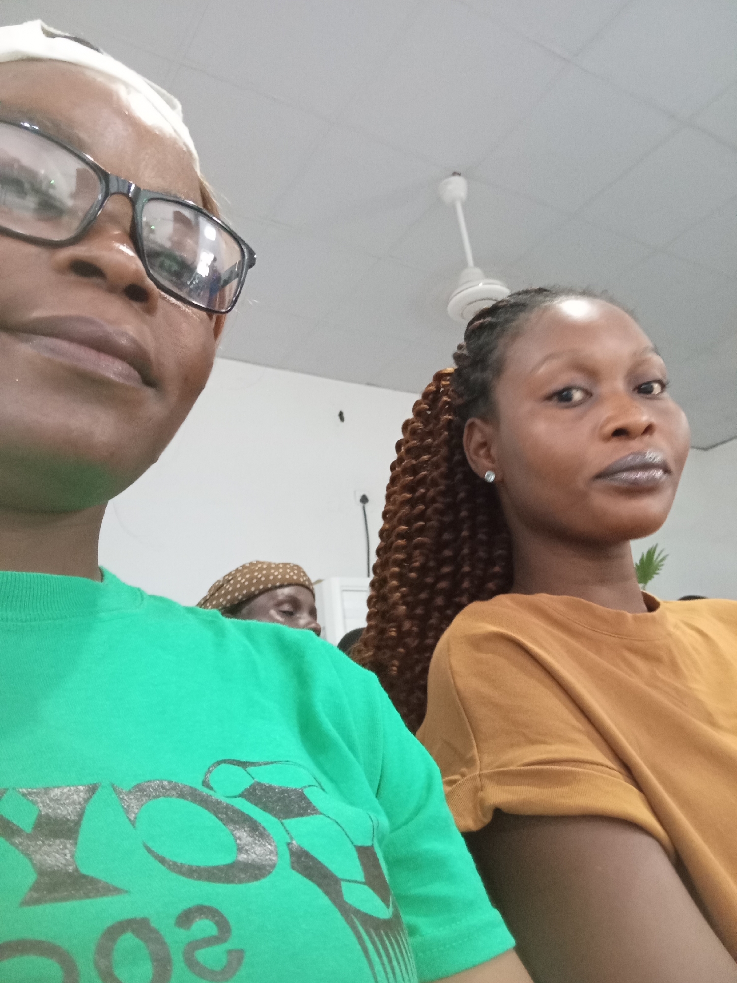 The image shows a close-up of two women, likely in an indoor setting. The woman on the left is wearing glasses and a green t-shirt with a graphic design. Her head is tilted, and she is looking slightly to the left. The woman on the right has braided hair and is wearing an orange t-shirt. She is looking directly at the camera. In the background, another person is partially visible, wearing a patterned head covering. A ceiling fan is visible in the upper portion of the image, suggesting the location is a room with a ceiling. The overall lighting is bright, indicating daytime or well-lit indoor conditions. The image does not contain any specific details that would allow for the identification of the city as Kason, Nigeria.