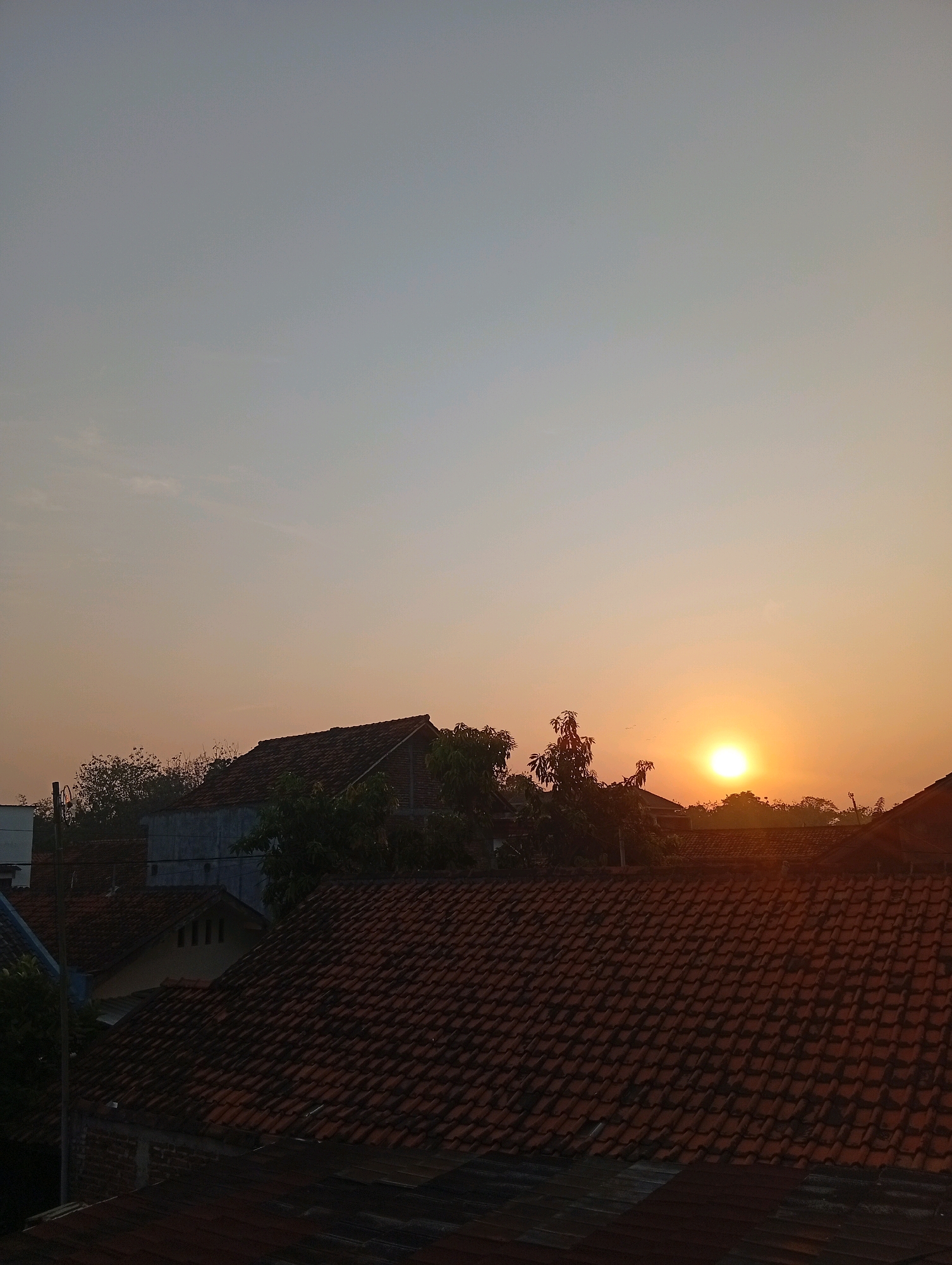 The image depicts a sunrise over a residential area in Kebandingan, Indonesia. The foreground is dominated by the tiled roofs of several houses, made of reddish-brown clay tiles. Some of these houses have visible brickwork or light-colored walls. In the middle ground, more houses and trees are visible, with their shapes softened by the morning haze. The sky above is a gradient of pale blue at the top, transitioning to a soft orange and yellow near the horizon, indicating the rising sun. The sun itself is a bright, circular orb, positioned slightly to the right of the frame, casting a warm glow. The overall atmosphere is peaceful and serene, characteristic of an early morning in a semi-urban or rural Indonesian setting. No people are visible in the image.