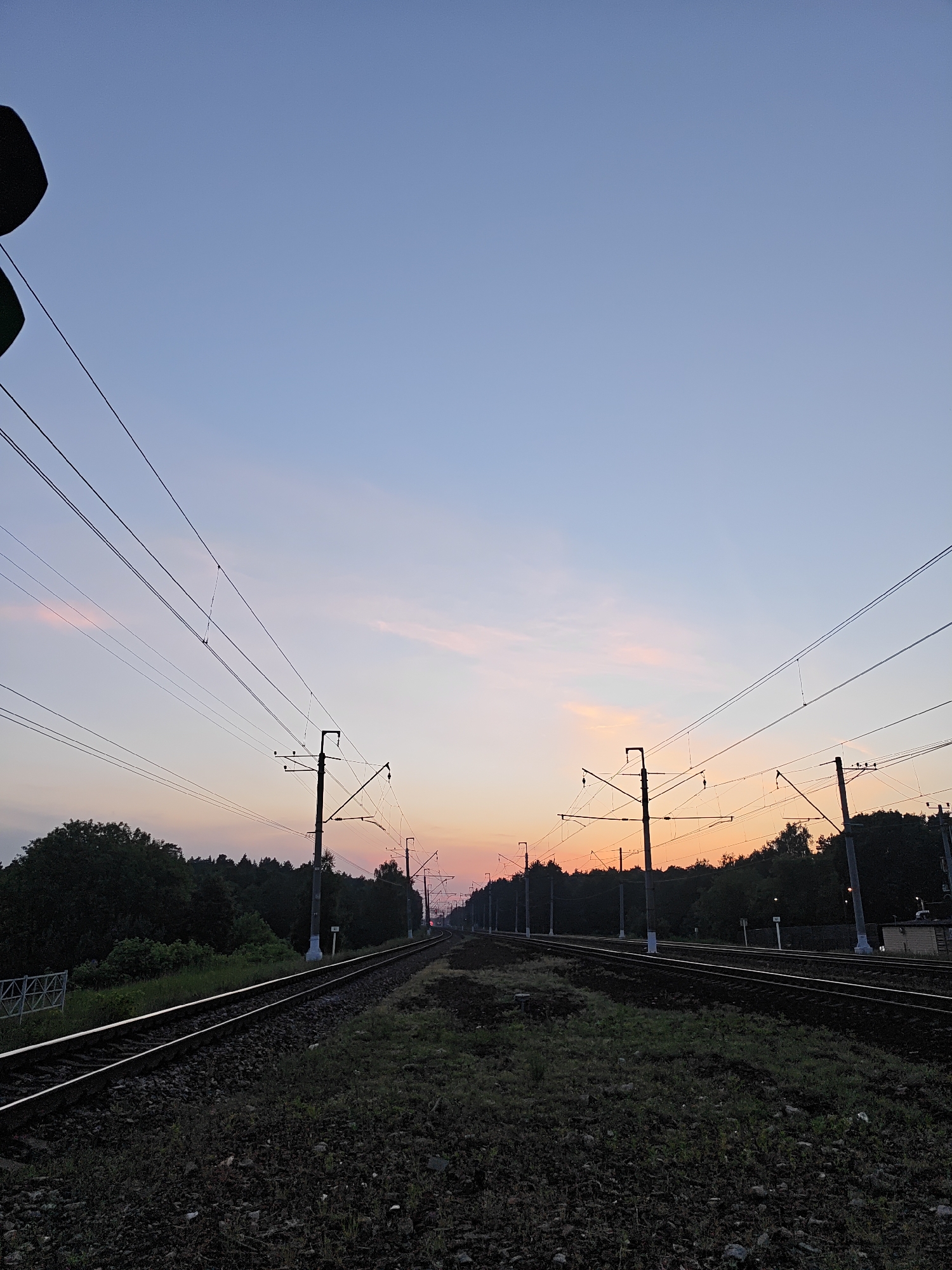 The image depicts a multi-track railway line in Kratovo, Russia, during twilight. The sky transitions from light blue at the zenith to pale pink and orange-red at the horizon, with sparse, thin clouds. Two main sets of parallel tracks, constructed on ballast, extend from the foreground into the distant curve. A dense network of overhead power lines is suspended by numerous dark utility poles flanking both sides of the tracks. Dark, deciduous forest lines the railway corridor on both sides, silhouetted against the sky. In the foreground, between the leftmost tracks, a white metal barrier is visible. Between the main sets of tracks, a white rectangular marker displaying "110" stands upright. In the far distance on the rightmost track, a faint red light source is discernible. The immediate foreground features dark ground cover, including ballast, dirt, and sparse vegetation. A partial view of a green signal or light fixture is present in the upper left corner of the frame. No individuals are present.