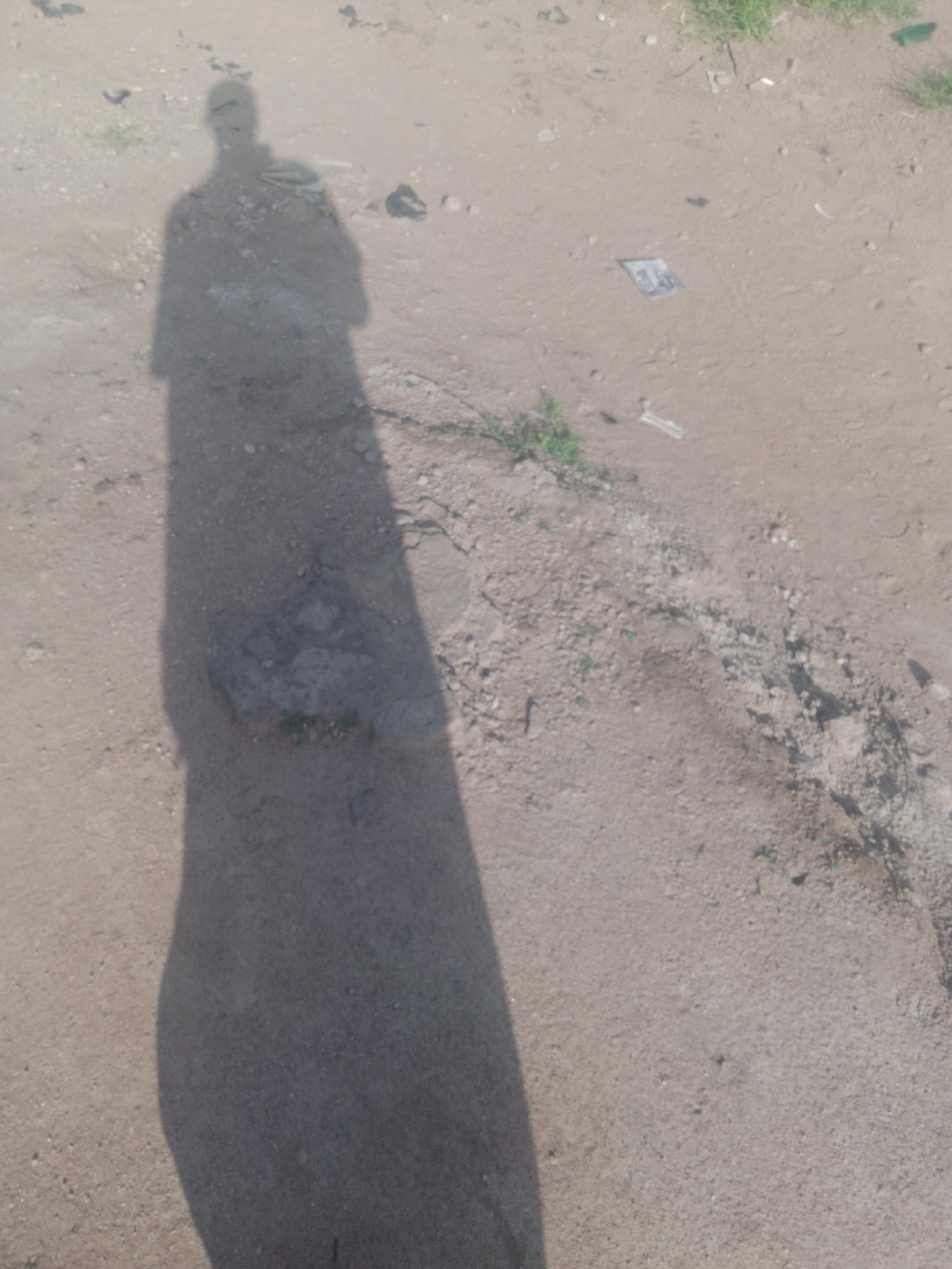 The image is a photograph of a person's shadow cast on a dirt surface. The shadow is elongated, suggesting the sun is low in the sky. The ground is composed of dirt, small stones, and patches of sparse vegetation. In the background, there are some scattered pieces of debris, including what appears to be a small, crumpled piece of paper or plastic. The scene is outdoors and appears to be in an undeveloped or unpaved area within the city of Jauro Yinu, Nigeria.