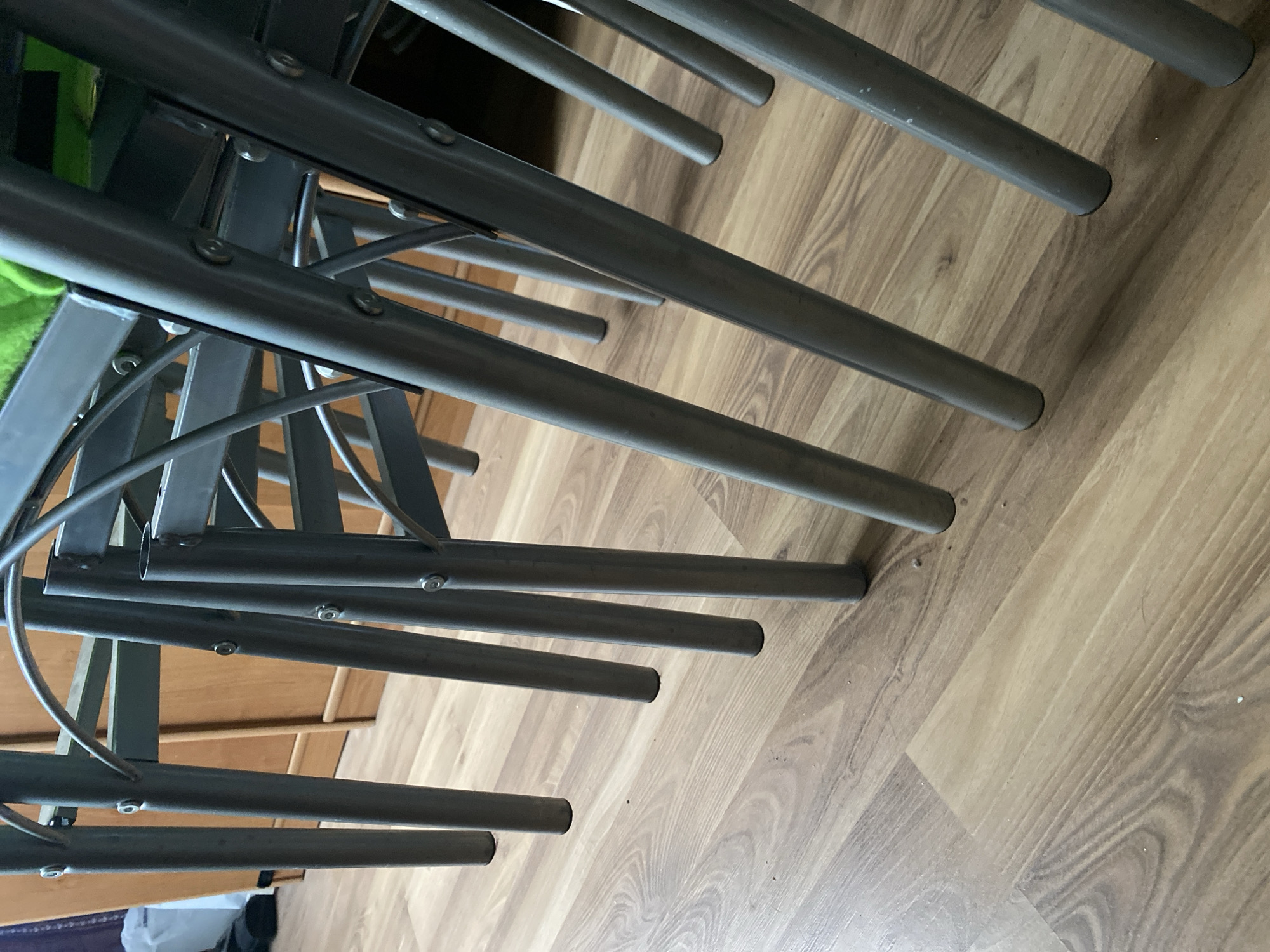 The image shows the legs of several metal chairs on a wooden floor. The chairs are stacked or closely positioned, with their grey metal legs extending downwards. The floor is made of laminate flooring with a wood grain pattern. In the background, a wooden cabinet or furniture piece is visible, along with a white plastic bag and some dark clothing on the floor to the left. The scene appears to be indoors, possibly in a home or storage area. There are no people or specific identifiable landmarks within the frame to suggest a location in Bílina, Czechia.