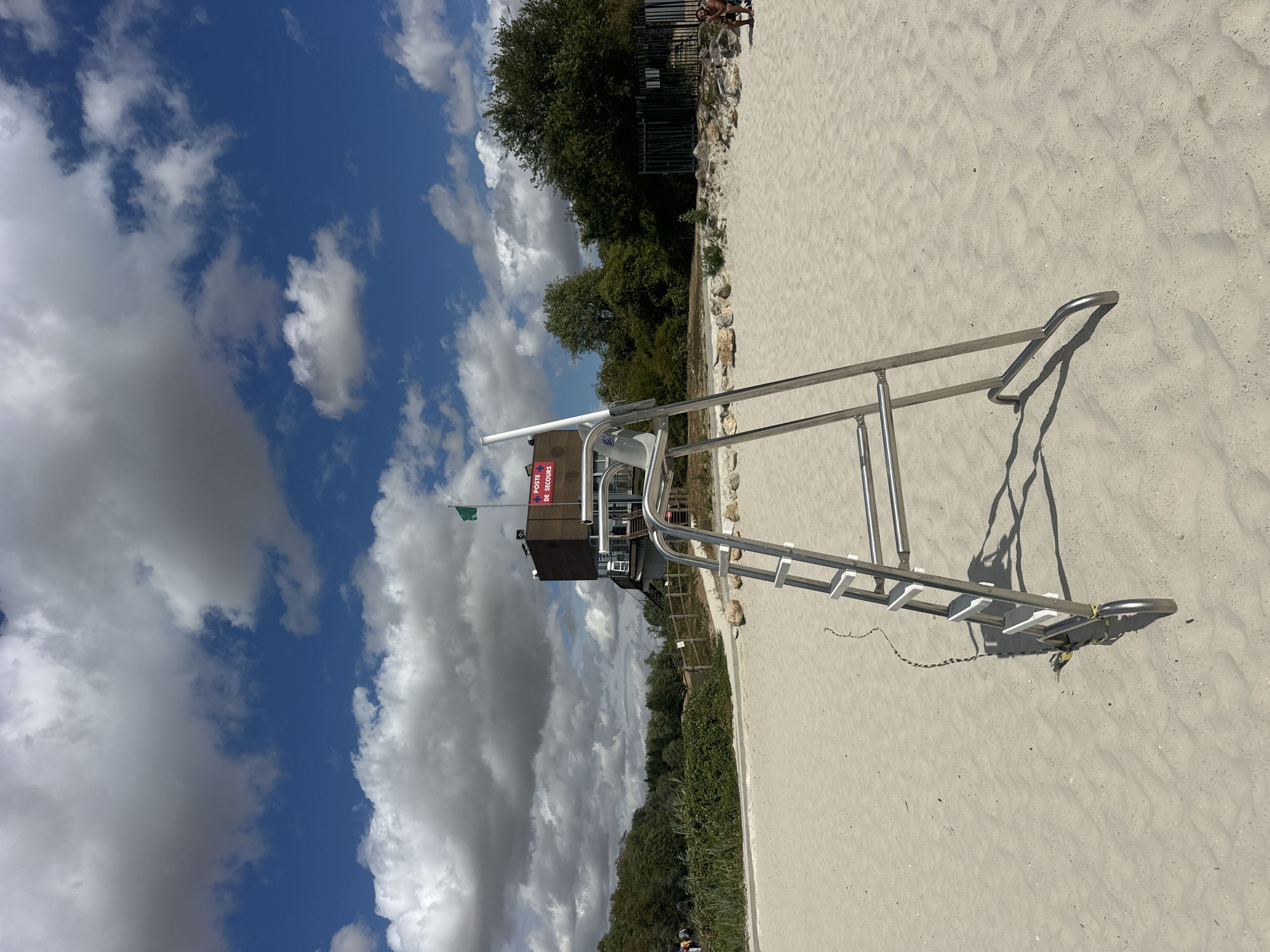 A lifeguard chair stands on a sandy beach in Jablines, France, beneath a blue sky with scattered white clouds. A building with a red cross emblem and French text reading "POSTE DE SECOURS" is visible in the background, along with a green flag on a pole. Trees and foliage line the edge of the beach. To the right, a person is seated on the sand. The scene is set during the daytime.